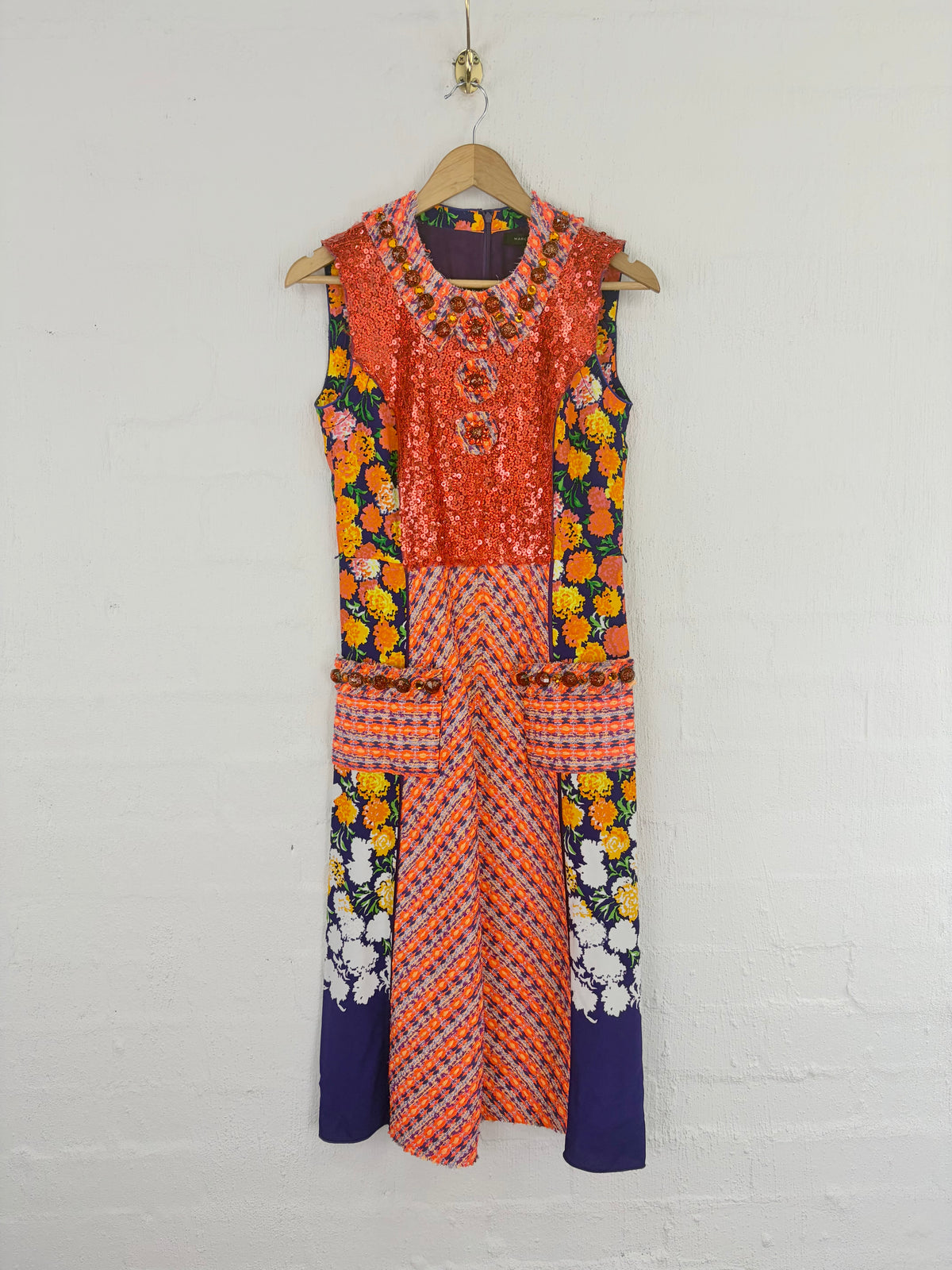 A sleeveless, multi-patterned dress hanging on a wooden hanger against a white brick wall. The dress features a bright orange and yellow floral pattern on the upper bodice and sleeves, a solid orange and red striped patterned waist section, and dark purple lower panels with white floral accents.