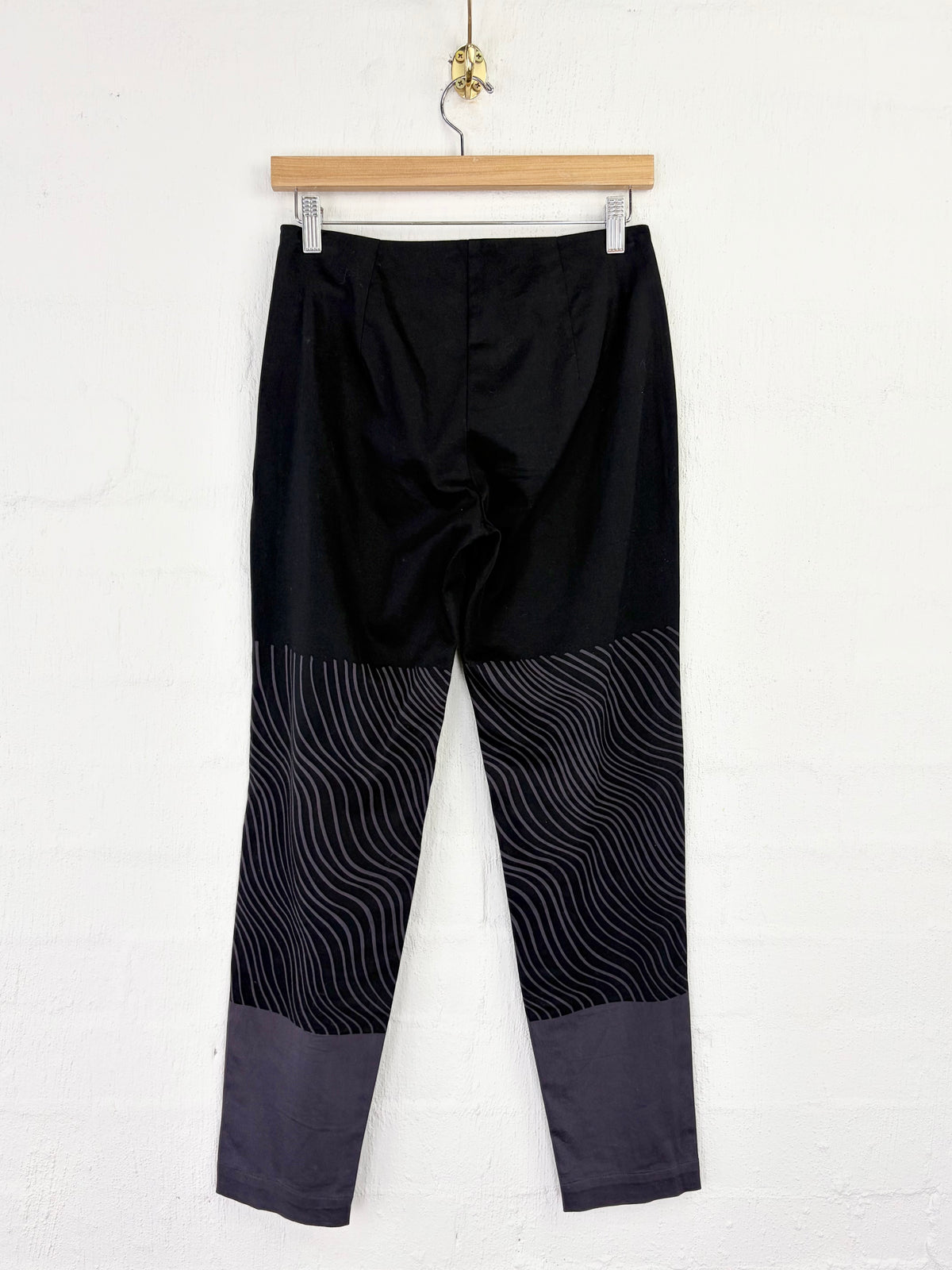Black cropped pants with a wavy patterned lower leg panel and dark gray cuffs, hanging from a wooden hanger against a white wall.