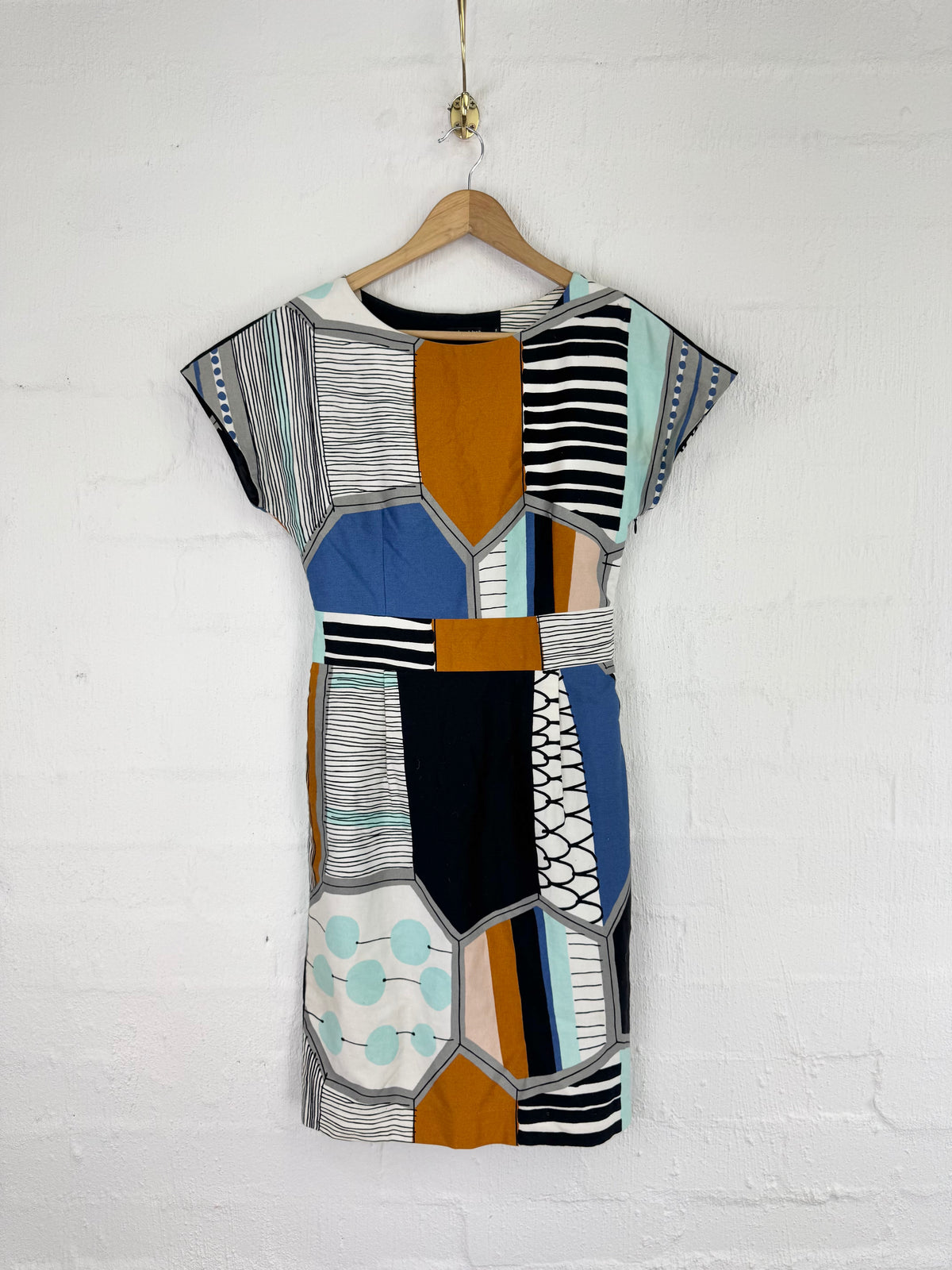 A short-sleeve dress with a geometric patchwork print in blue, orange, black, white, and gray, hanging on a wooden hanger against a white brick wall.