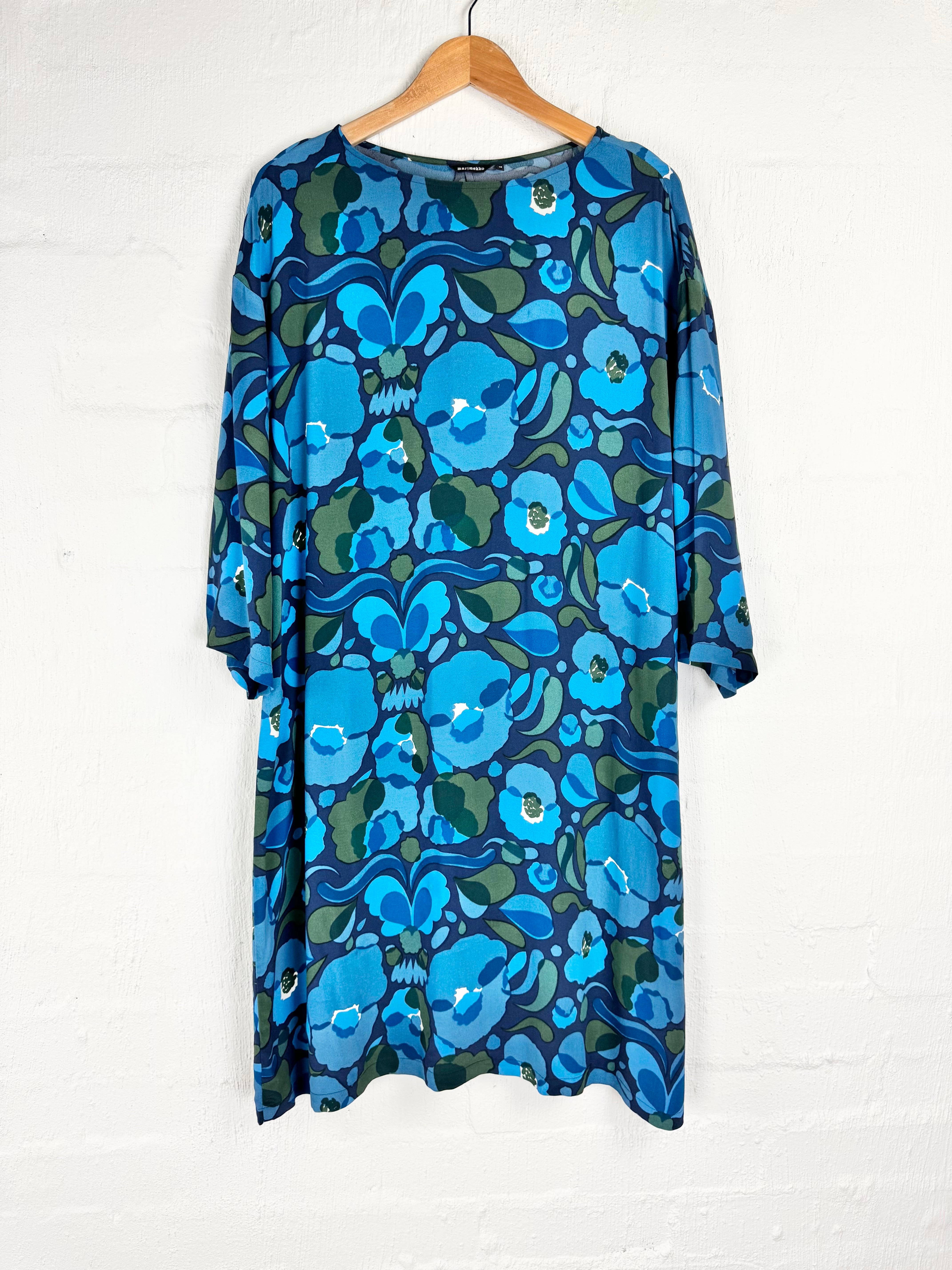 A blue and green floral print dress with three-quarter sleeves hanging on a wooden hanger against a white brick wall.