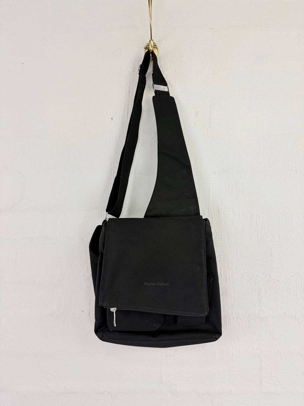 A black fabric shoulder bag with a wide adjustable strap, hanging from a gold hook against a white textured wall. The bag features a front flap pocket with a small white logo and a zippered bottom compartment.