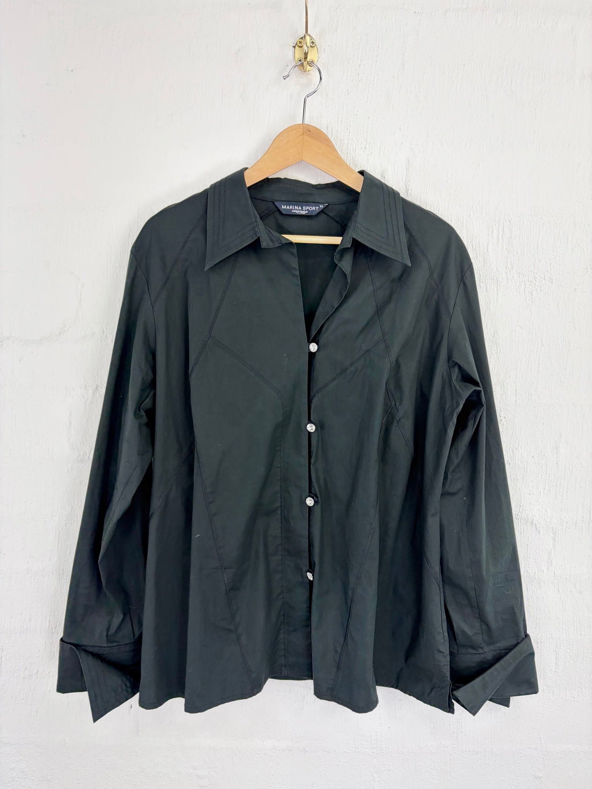 A black long-sleeve button-up shirt with a pointed collar, hanging on a wooden hanger against a white textured wall.