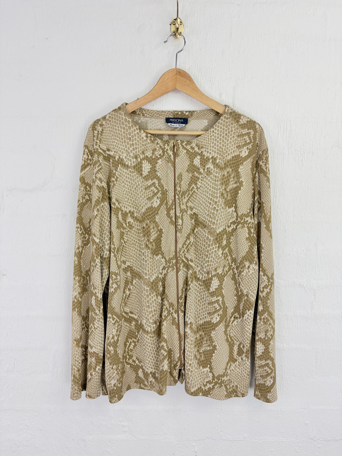 A beige and brown snakeskin print long-sleeve top hanging on a wooden hanger against a white painted brick wall.