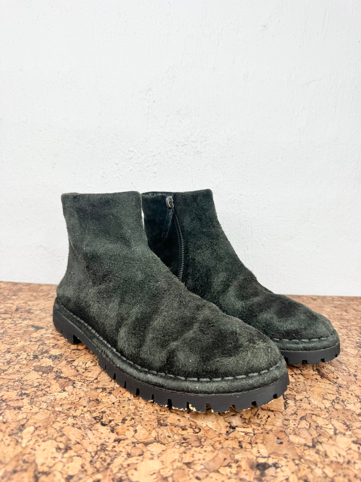 A pair of dark green suede ankle boots with black soles, displayed on a cork surface against a white wall.