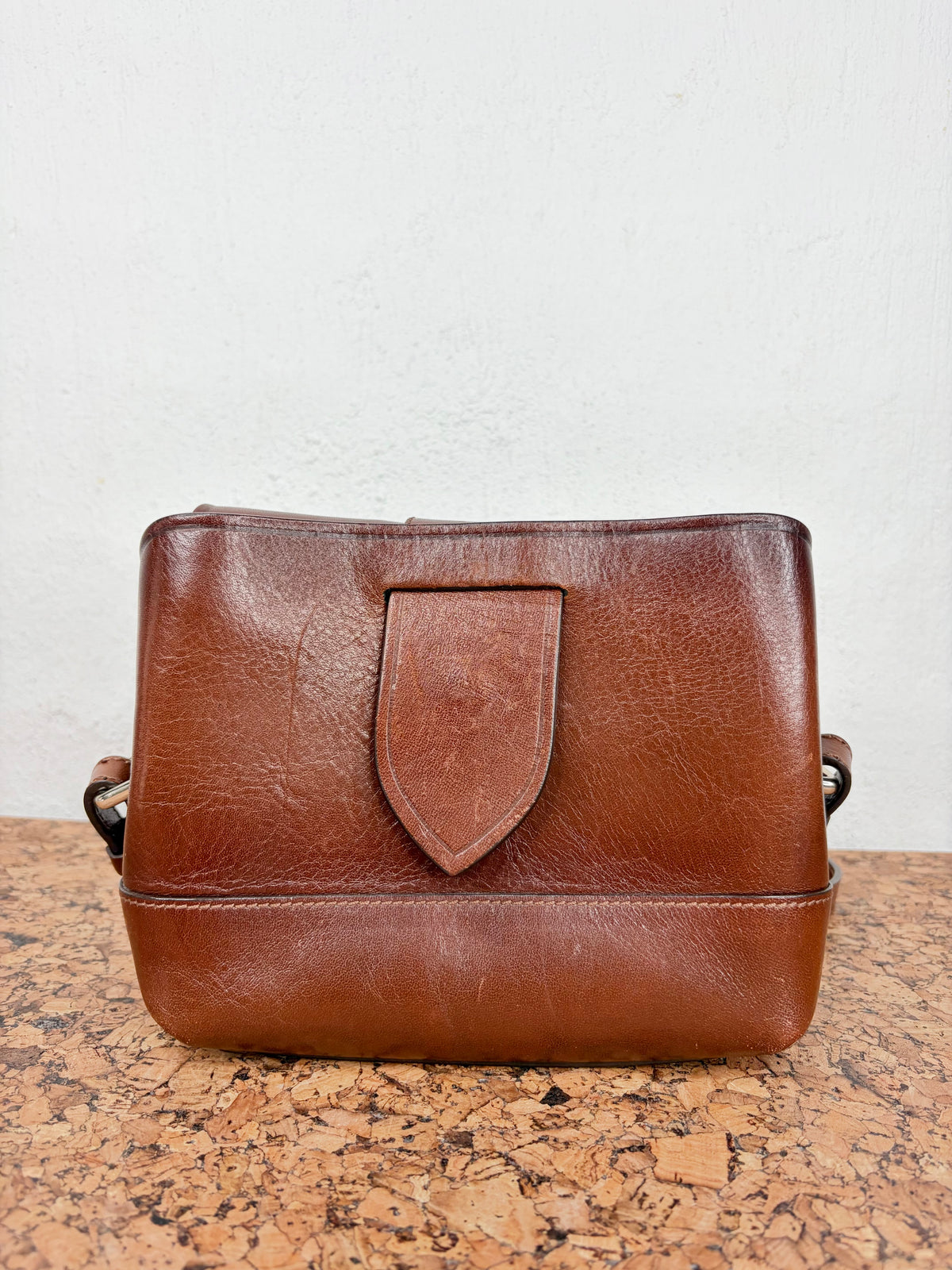 A brown leather shoulder bag with a shield-shaped front tab, resting on a cork surface against a white wall.