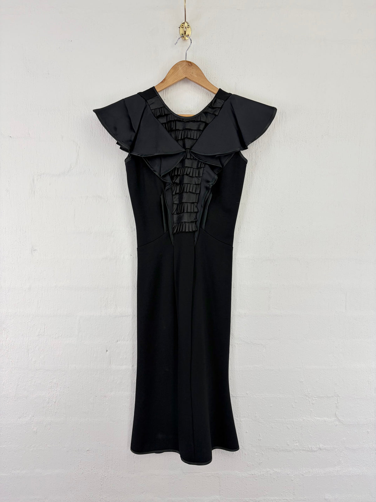 A black sleeveless dress with dramatic ruffled shoulder panels and a ruched smocked bodice, hanging on a wooden hanger against a white painted brick wall.