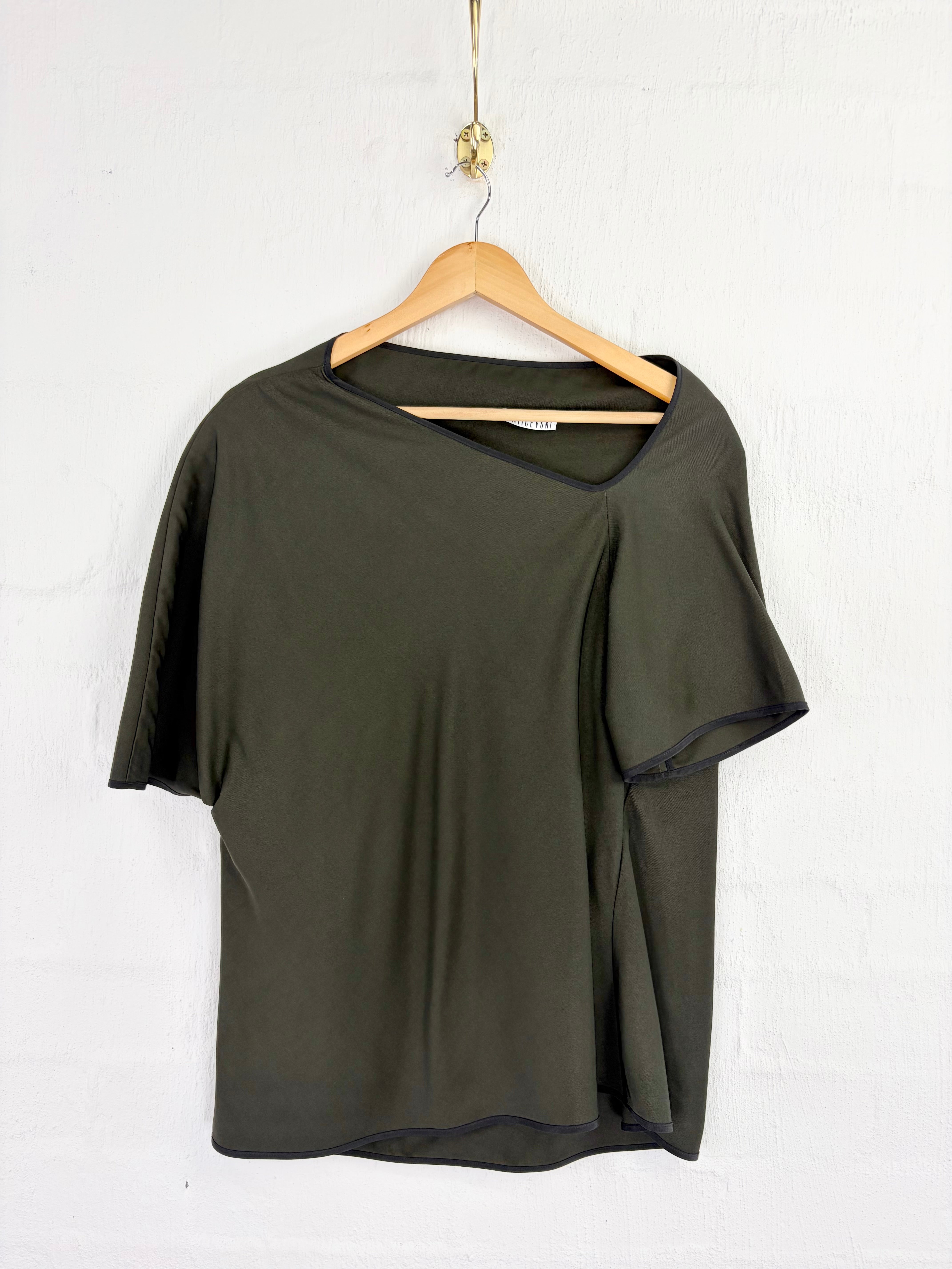 An olive green short-sleeve top with an asymmetrical draped sleeve detail, hanging on a wooden hanger against a white textured wall.