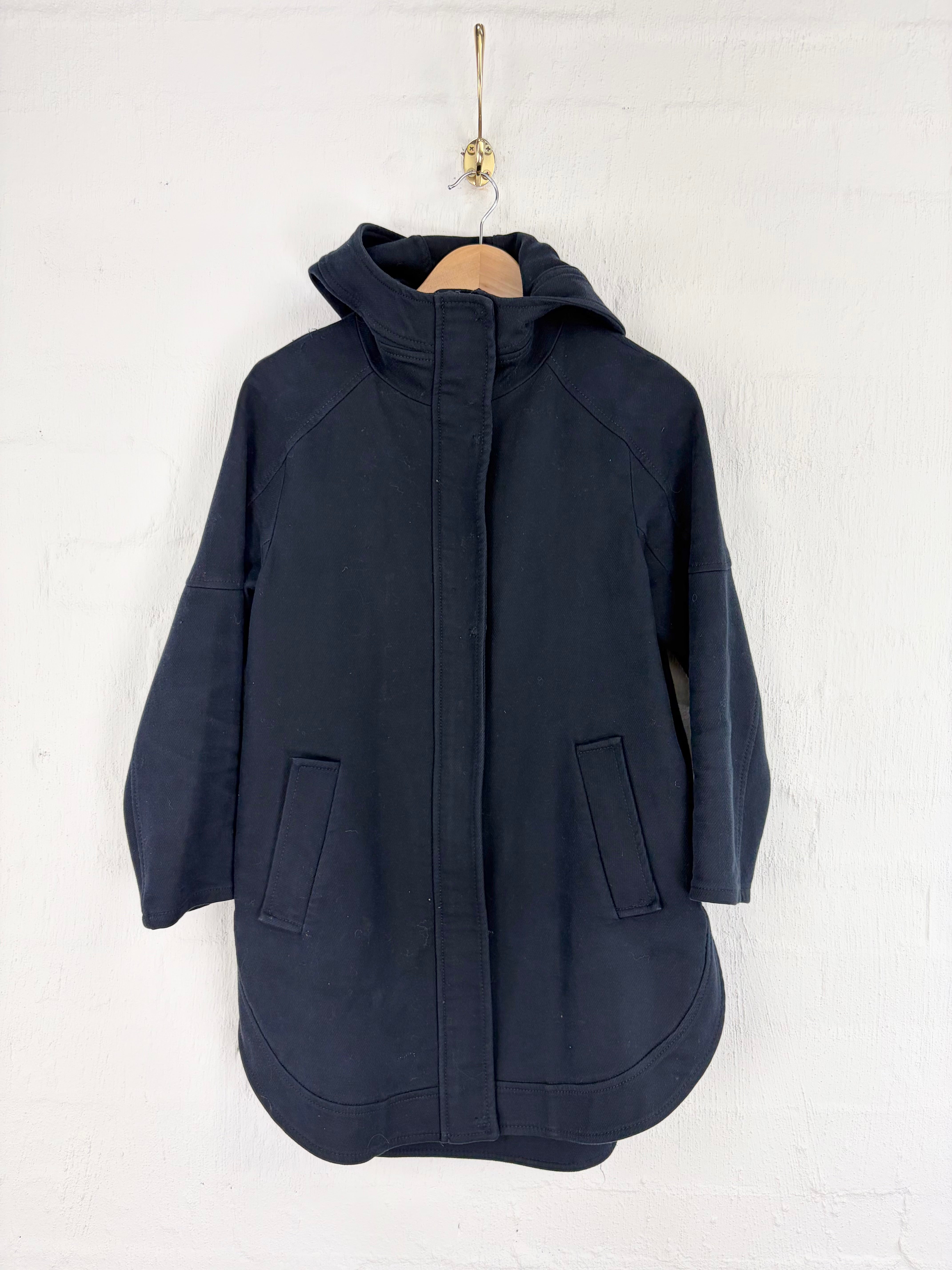 A dark navy hooded jacket with a front zipper and two large front patch pockets, hanging on a wooden hanger against a white painted brick wall.