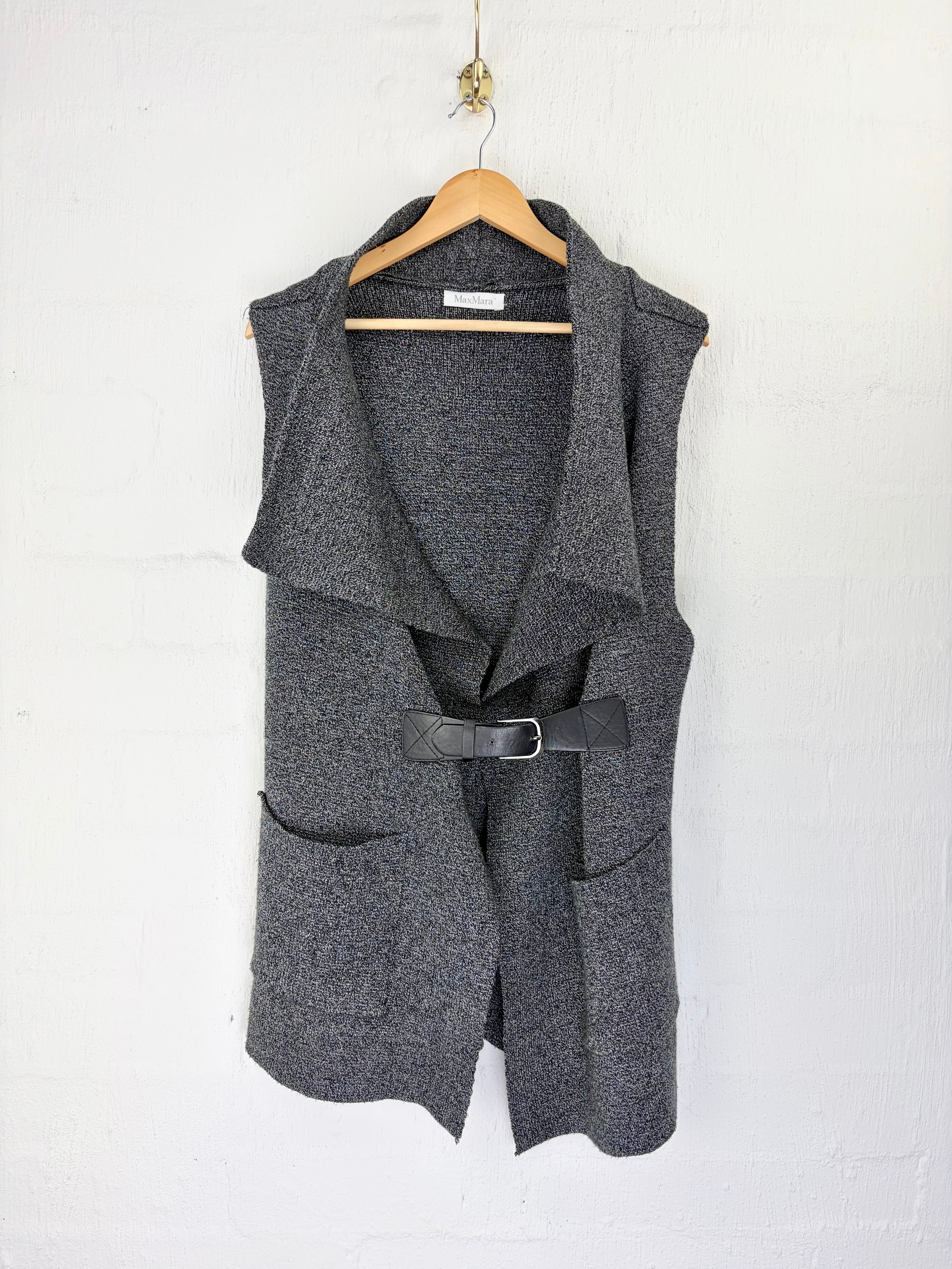 A dark gray sleeveless open-front vest with a black belt, hanging on a wooden hanger against a white painted brick wall.