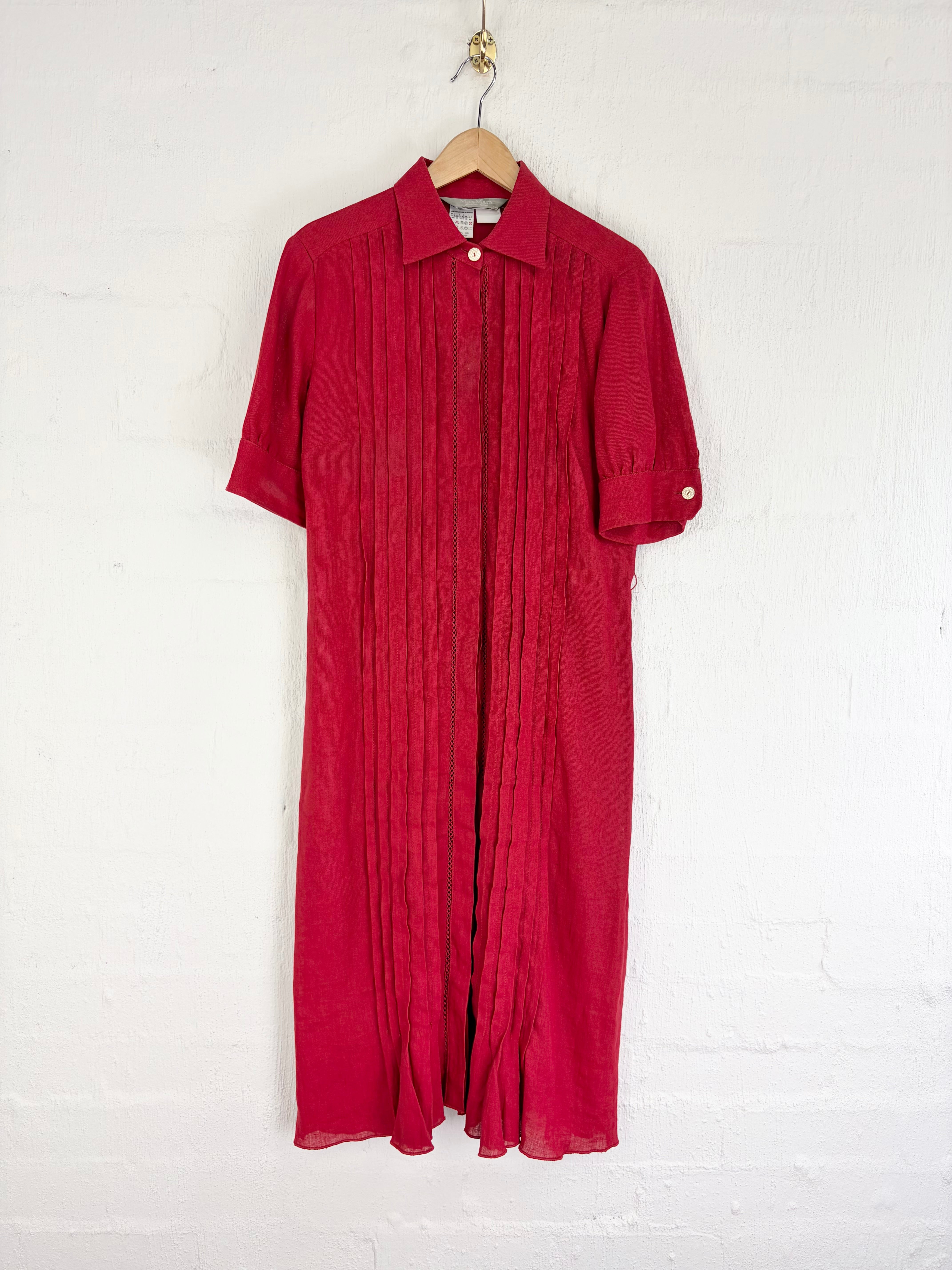 A red short-sleeve collared shirt dress with vertical pleating, hanging on a wooden hanger against a white painted brick wall.