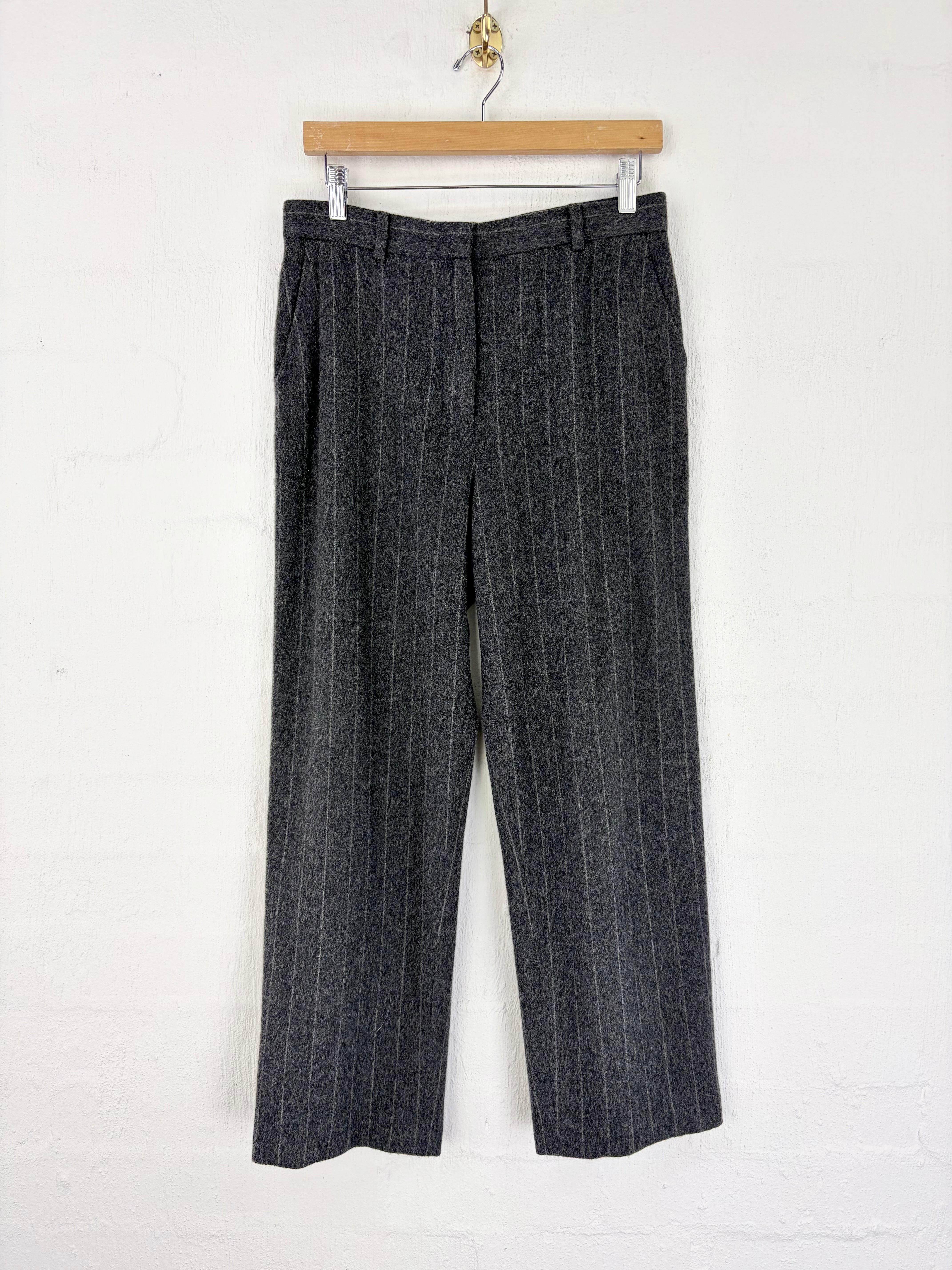 Dark gray pinstriped wool trousers hanging from a wooden hanger against a white painted brick wall.