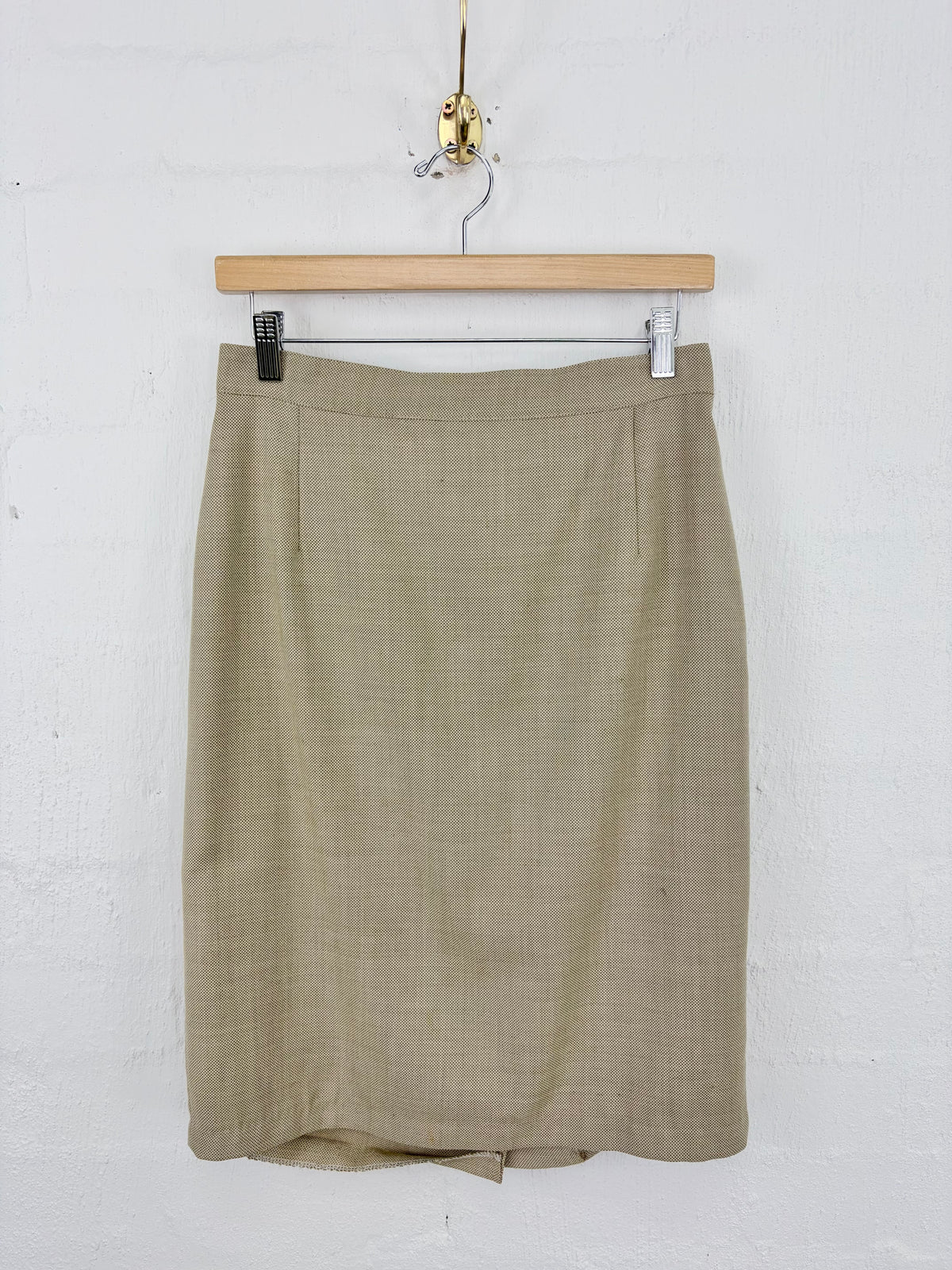 A knee-length olive green skirt with a subtle woven texture, hanging from a wooden hanger against a white painted brick wall.
