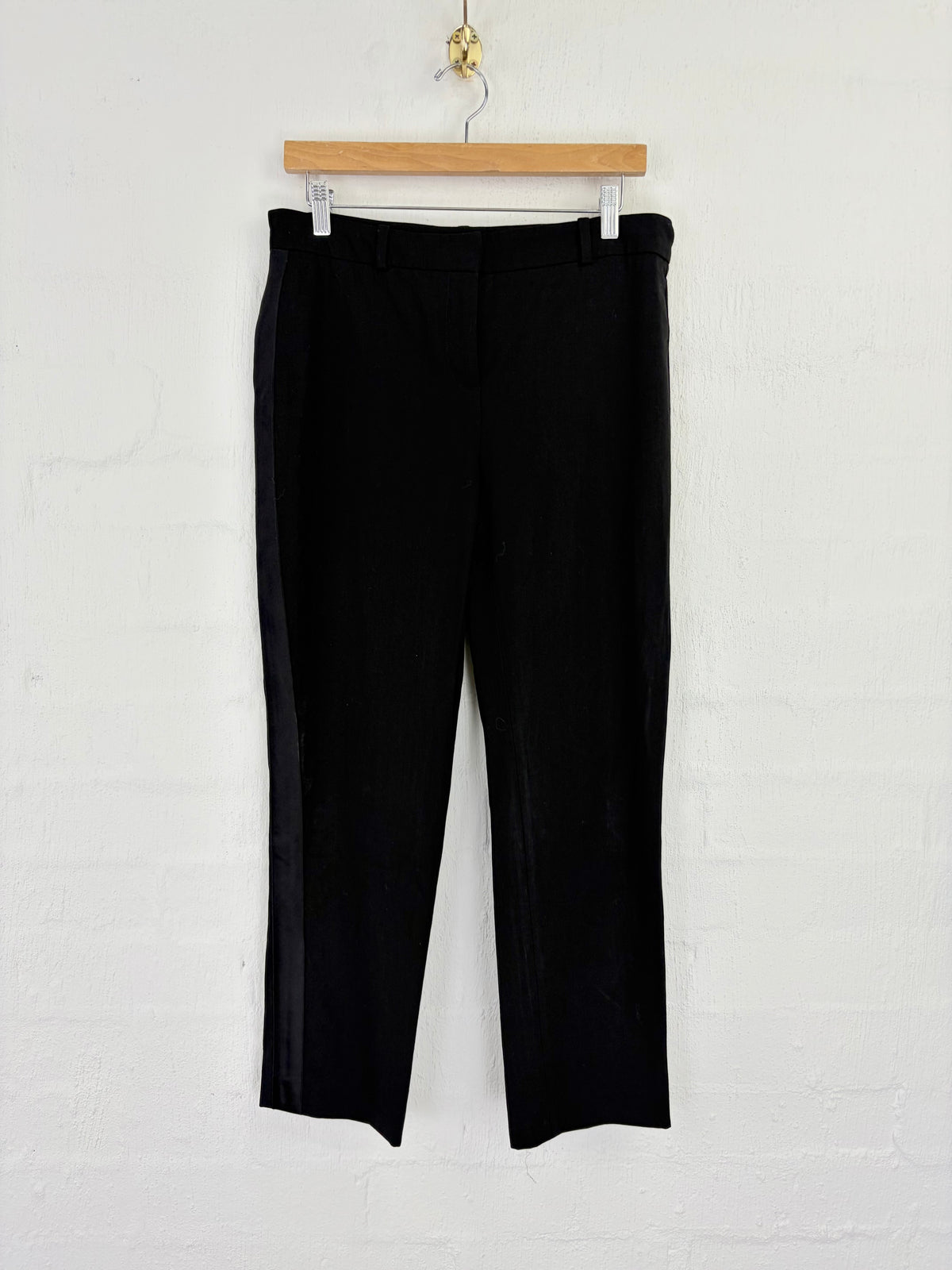 Black cropped trousers hanging from a wooden hanger against a white painted brick wall.