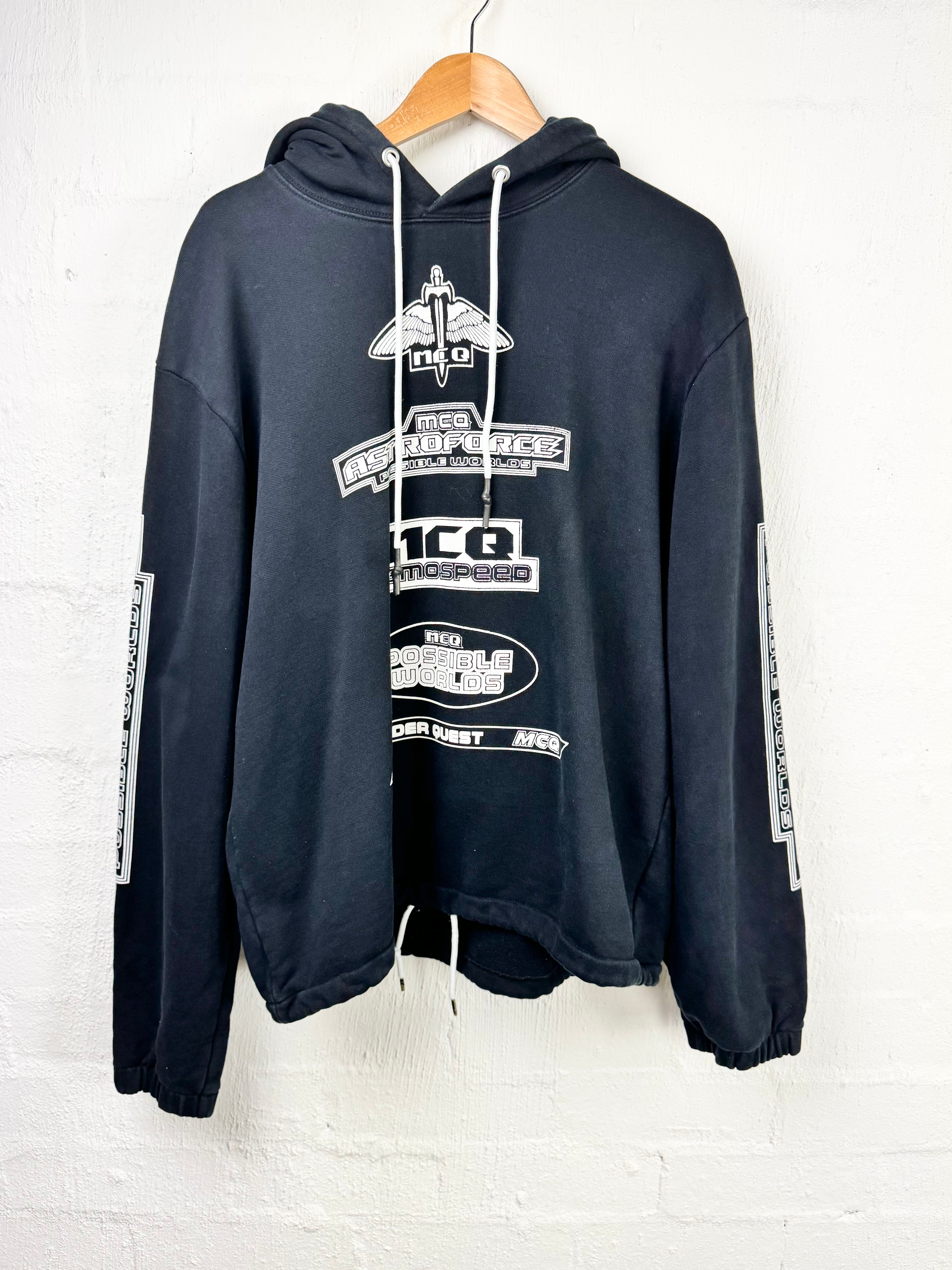 A dark navy blue hooded sweatshirt with white graphic prints and drawstrings, hanging on a wooden hanger against a white brick wall.