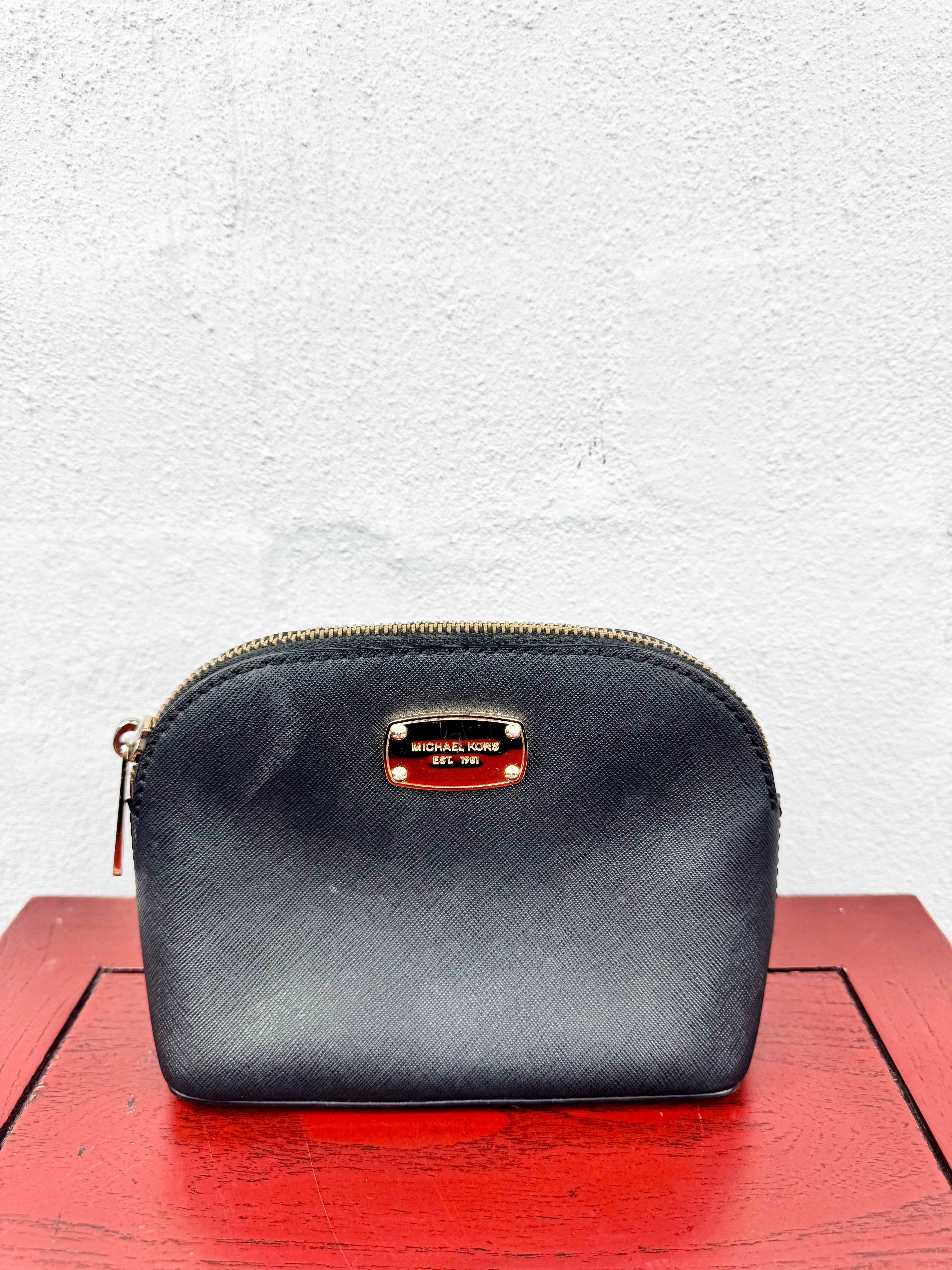 A Michael Kors black textured leather zippered pouch with a gold-tone metal logo plate, resting on a red wooden surface against a white textured wall.