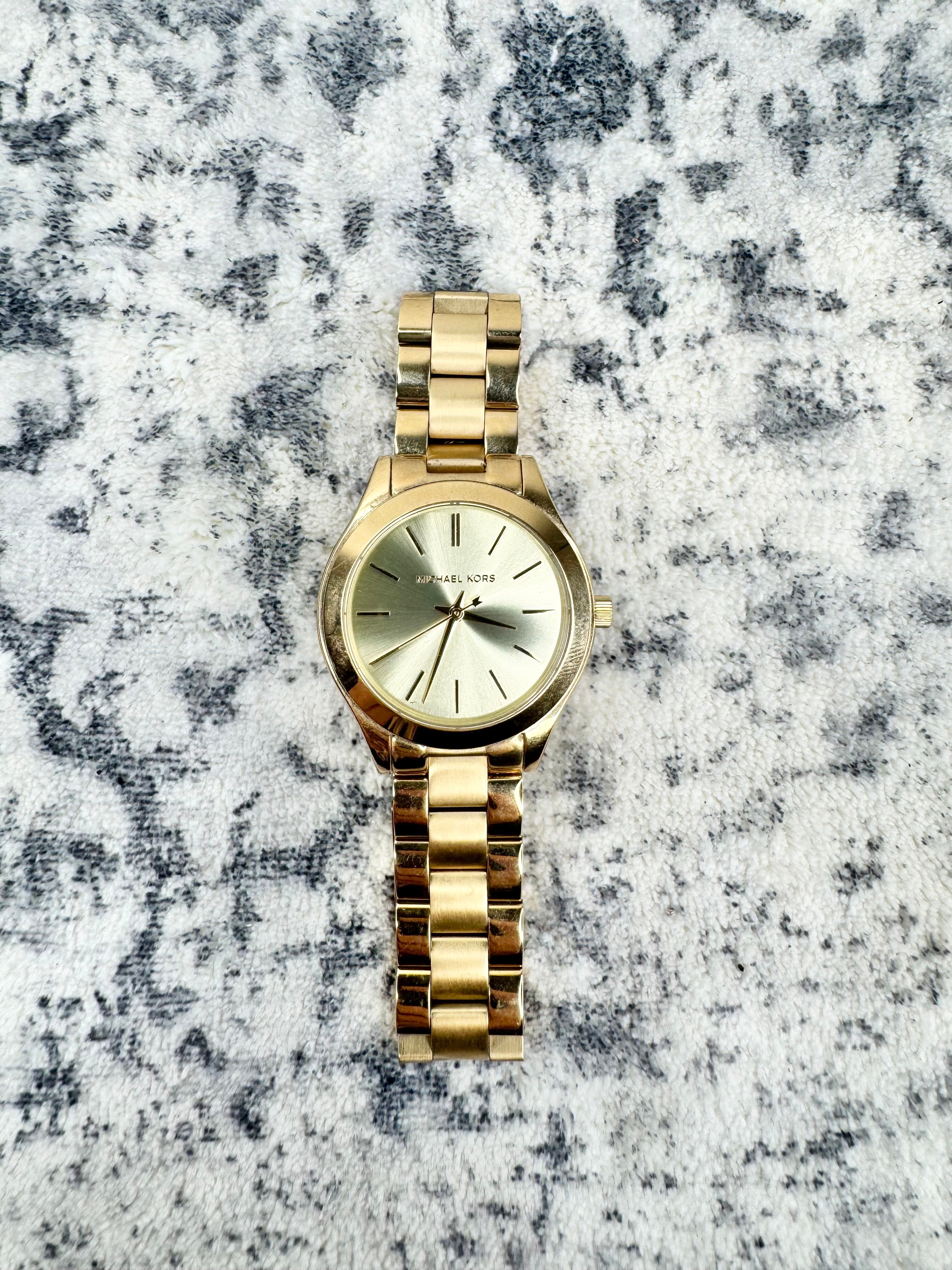 A gold-tone Michael Kors analog watch with a matching metal link bracelet and a light dial, placed on a textured gray and white surface.