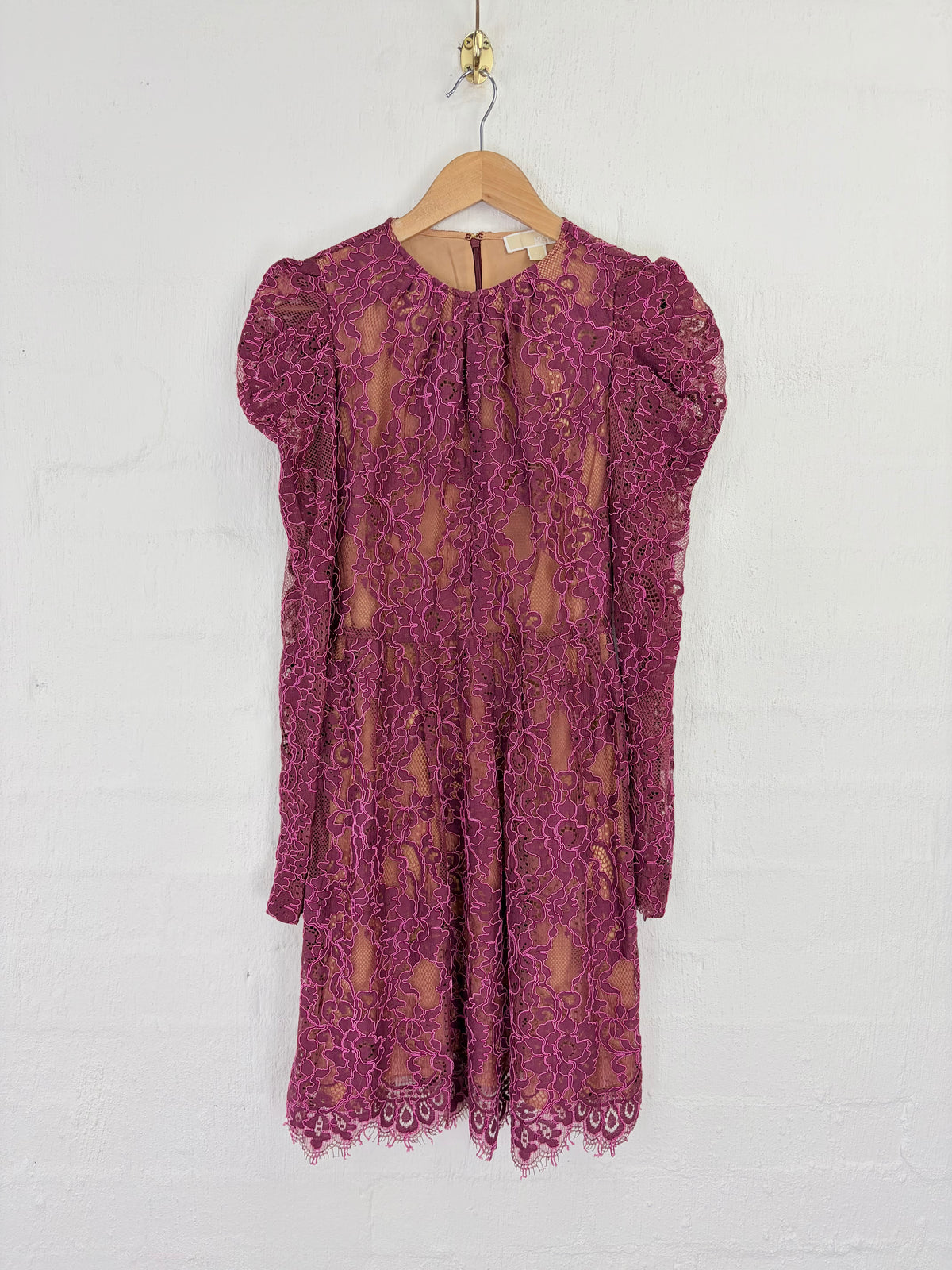 A magenta lace dress with short puff sleeves and a tie-neck detail, hanging on a wooden hanger against a white painted brick wall.