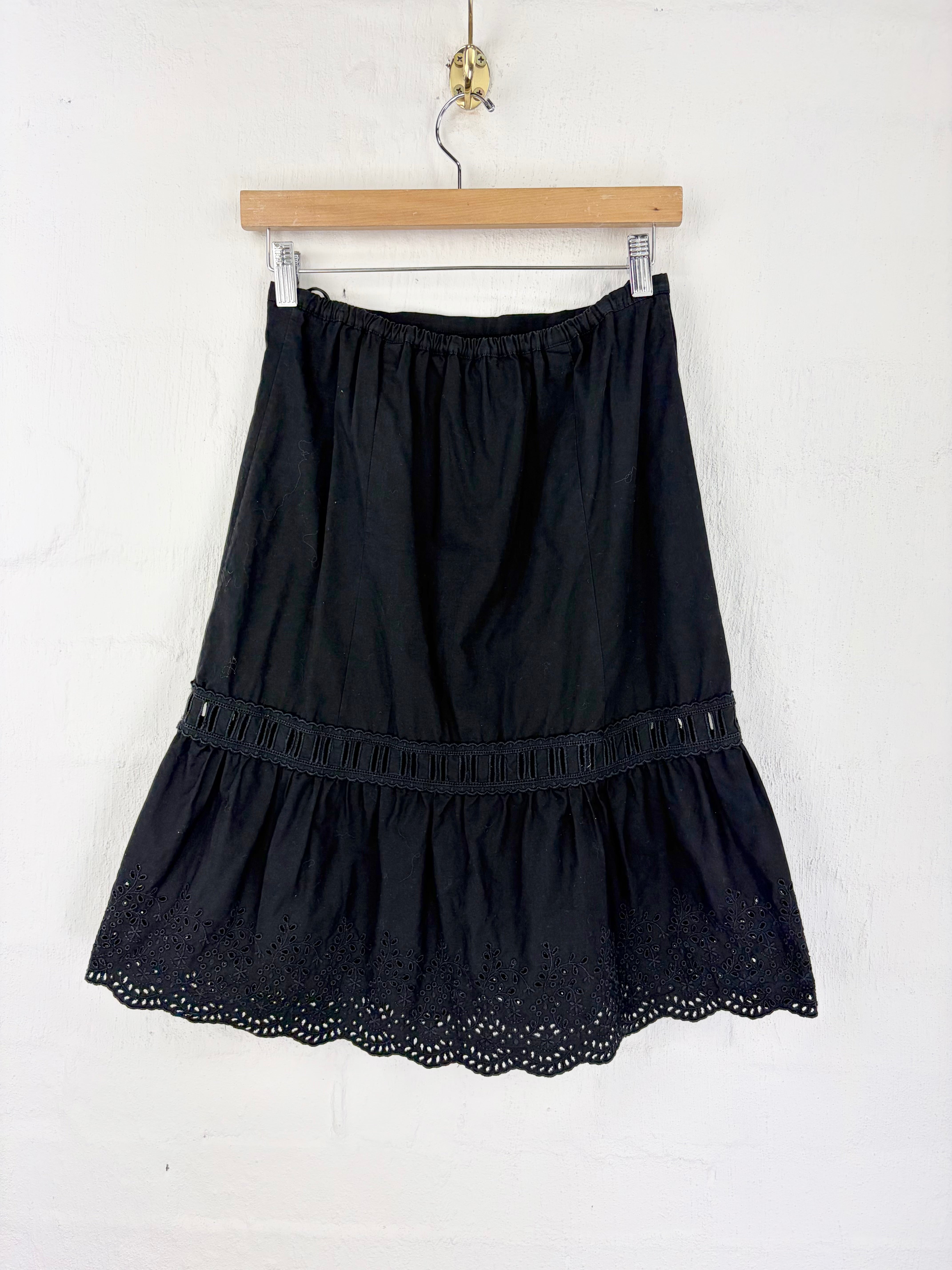 A black tiered skirt with eyelet lace trim and a scalloped hem, hanging from a wooden hanger against a white wall.