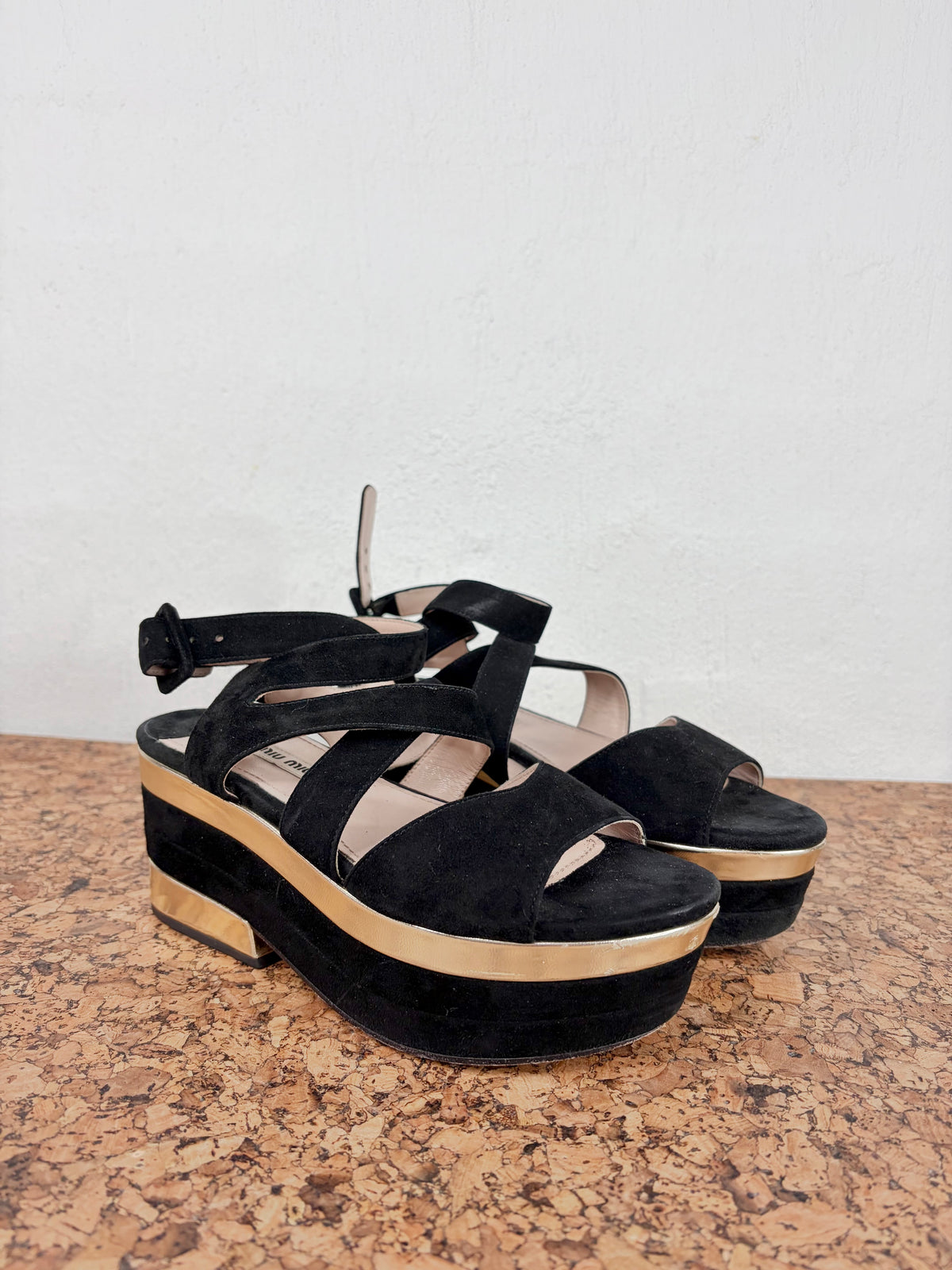 A pair of black platform wedge sandals with ankle straps and open toes, resting on a brown speckled surface against a white wall.