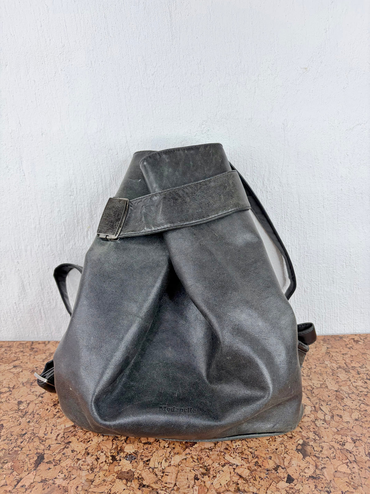 A dark gray distressed leather bucket bag with a strap detail, resting on a brown speckled surface against a white wall.