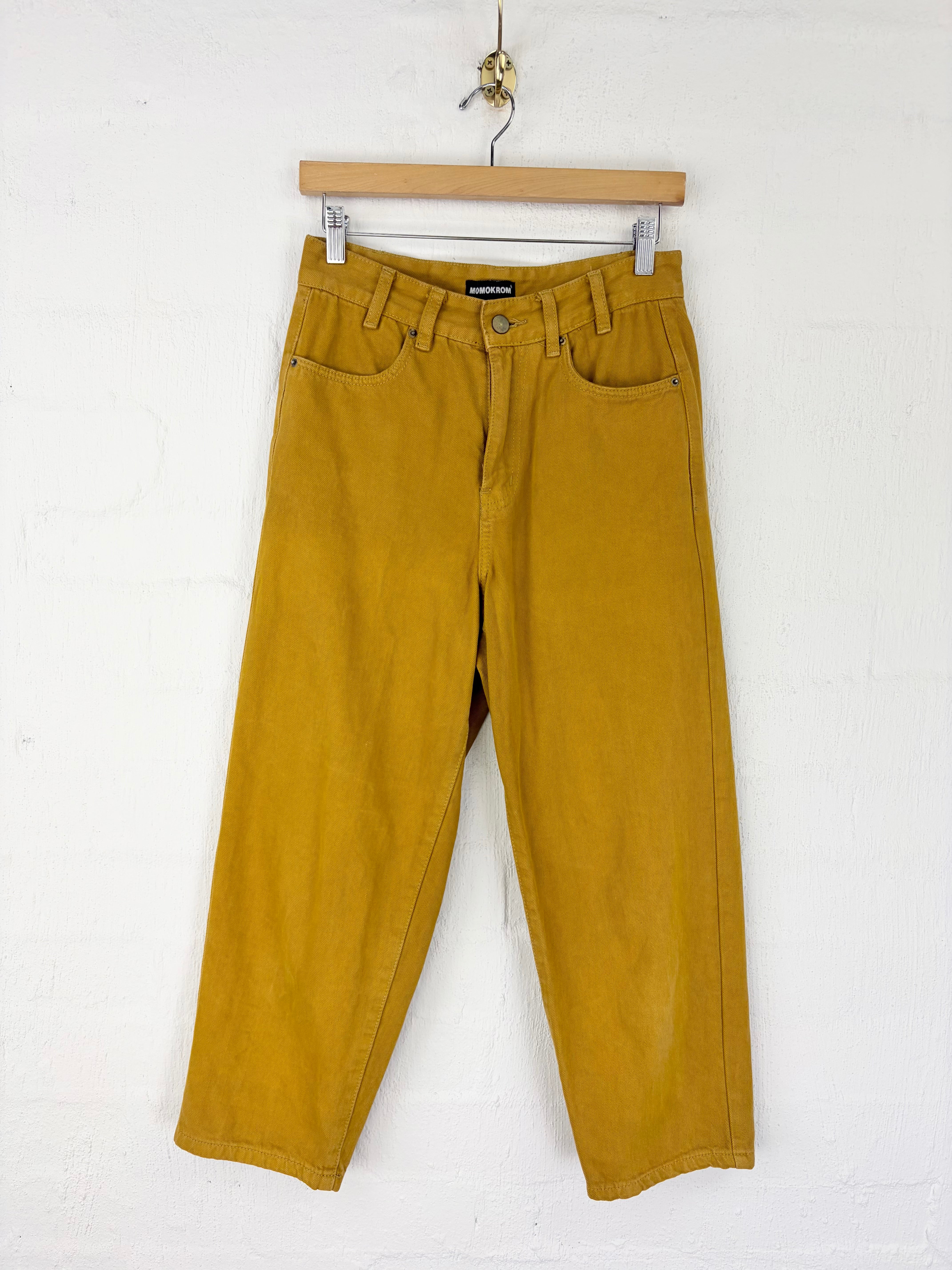 A pair of mustard yellow corduroy pants hanging from a wooden hanger against a white wall.