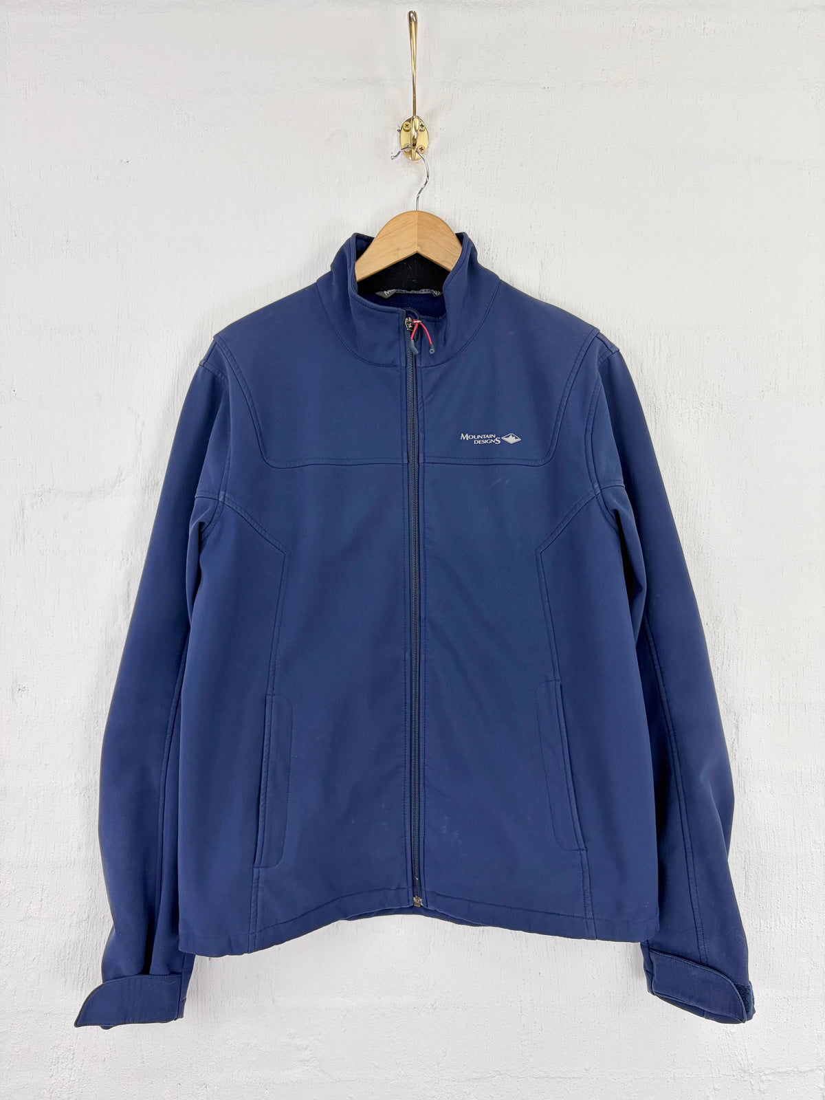 A navy blue full-zip jacket hanging on a wooden hanger against a white painted brick wall. The jacket features a small white embroidered logo on the left chest.