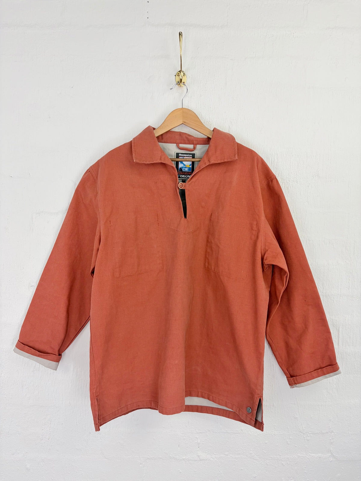 A terracotta-colored long-sleeve quarter-zip pullover shirt hanging on a wooden hanger against a white painted brick wall.