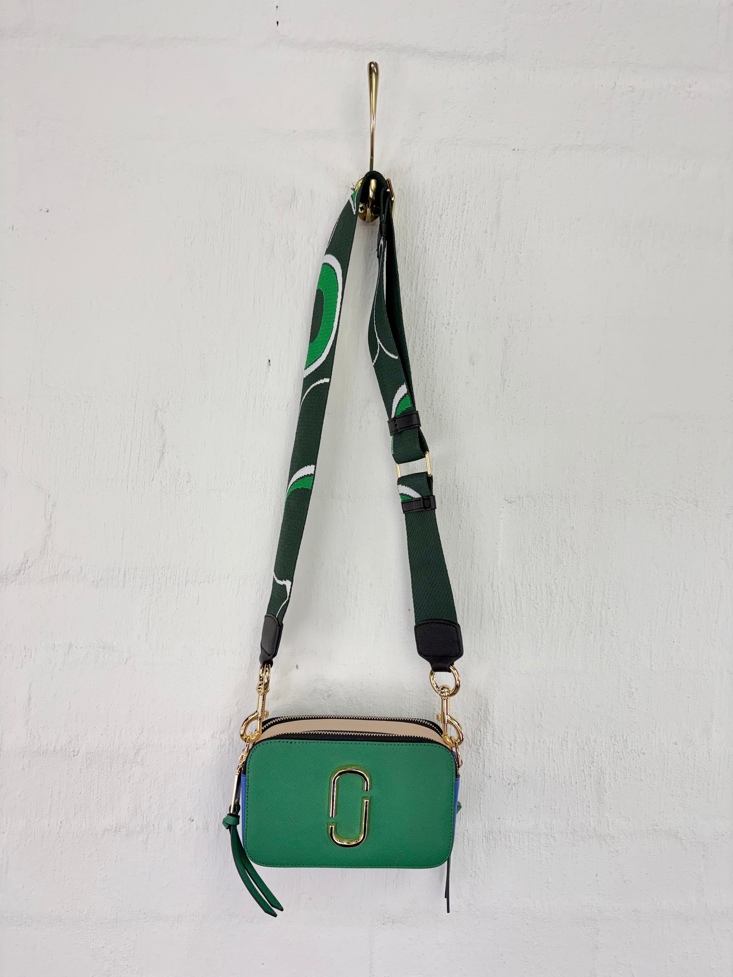A green crossbody camera bag with a patterned green and white strap, hanging from a gold hook on a white brick wall.