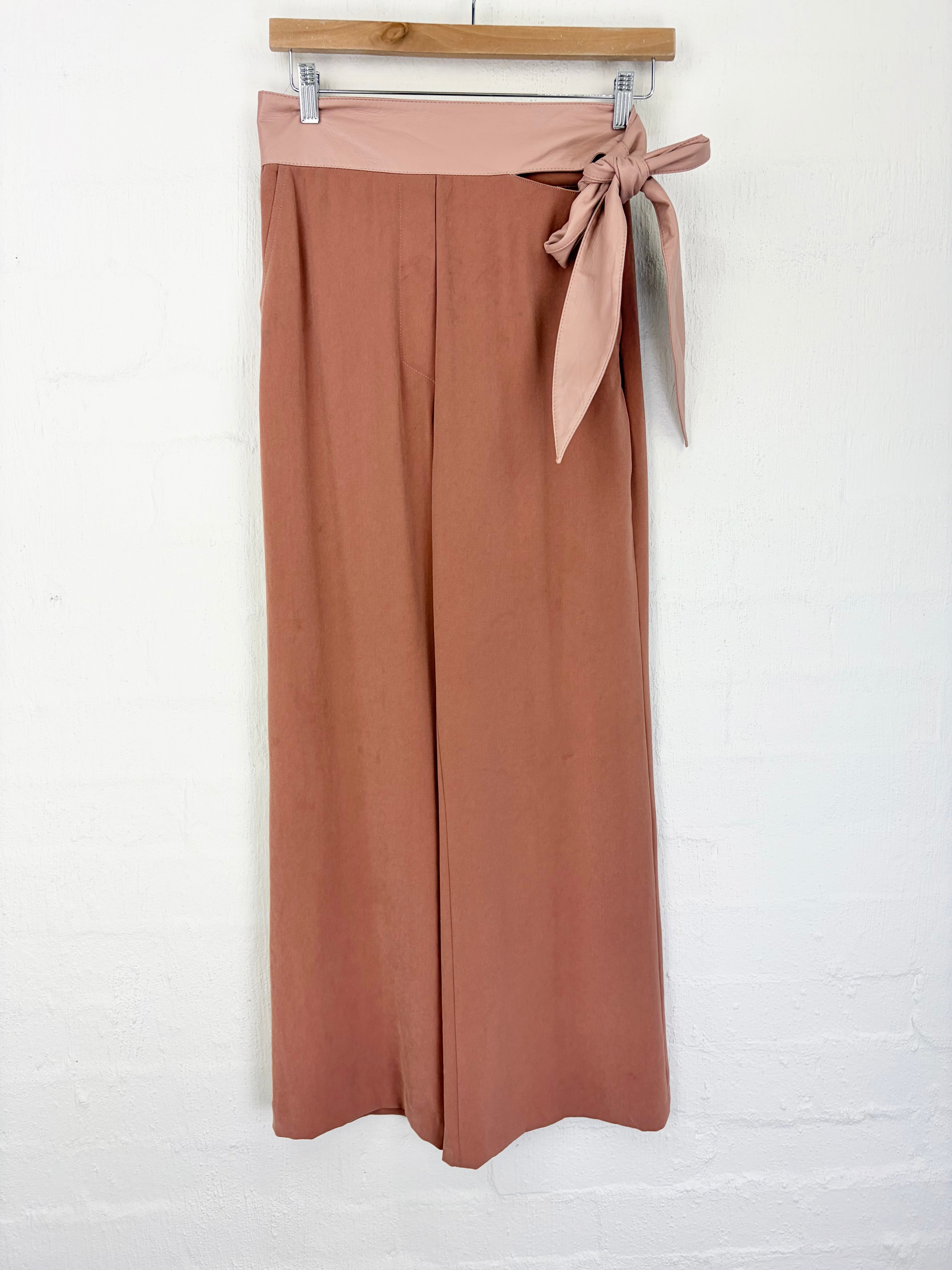 A pair of wide-leg cropped pants in a dusty rose-brown color, hanging from a wooden hanger against a white painted brick wall. The pants feature a matching fabric tie belt at the side waist.