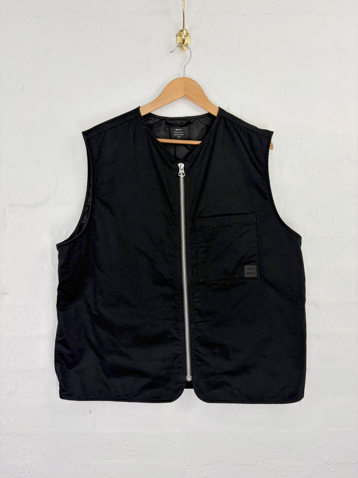 A black sleeveless zip-front vest hanging on a wooden hanger against a white textured wall. The vest has a full front zipper, a chest pocket with a small square label, and a visible interior neck label.