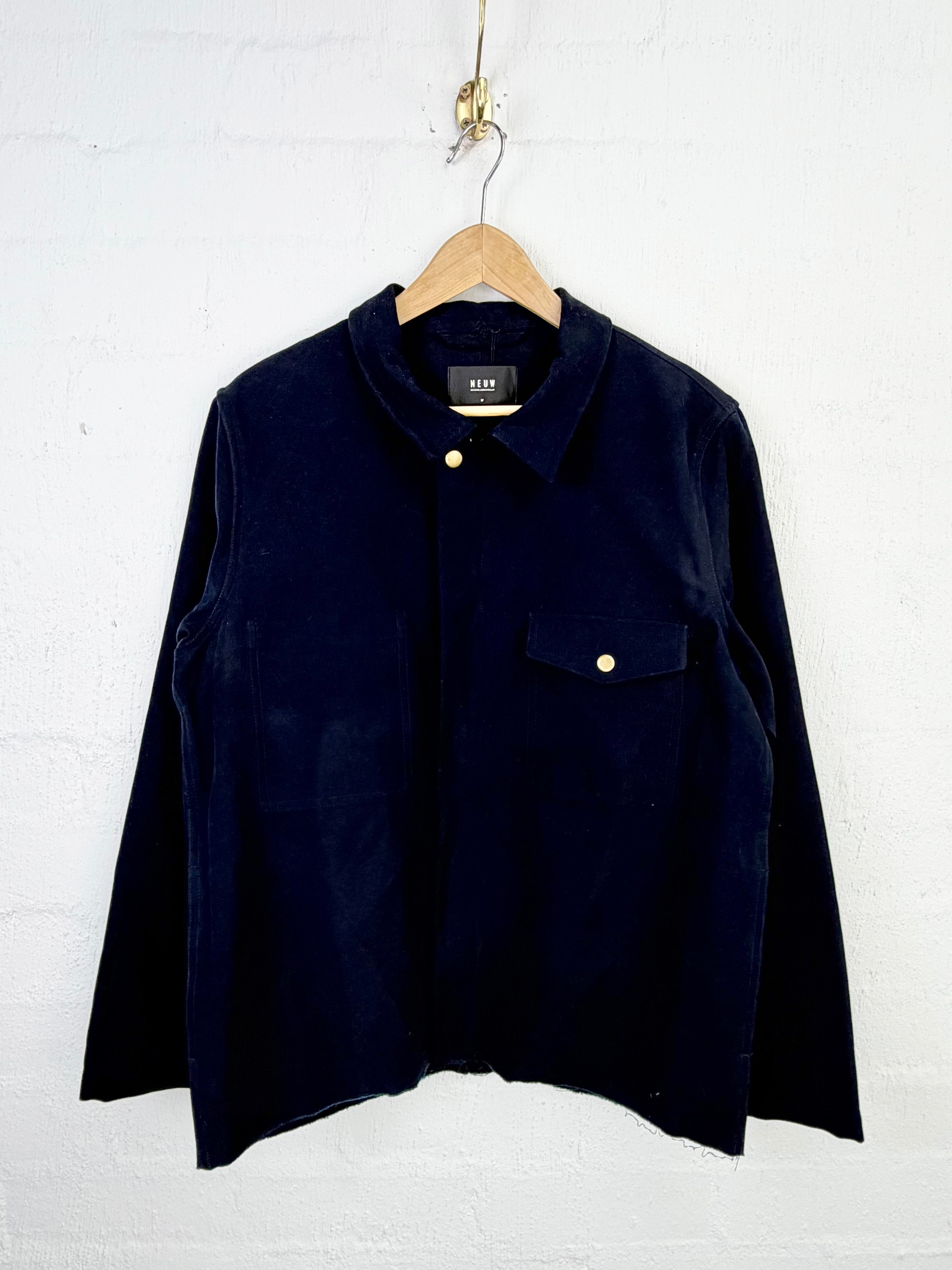 A dark navy blue corduroy shirt jacket with a pointed collar and a chest pocket, hanging on a wooden hanger against a white painted brick wall.