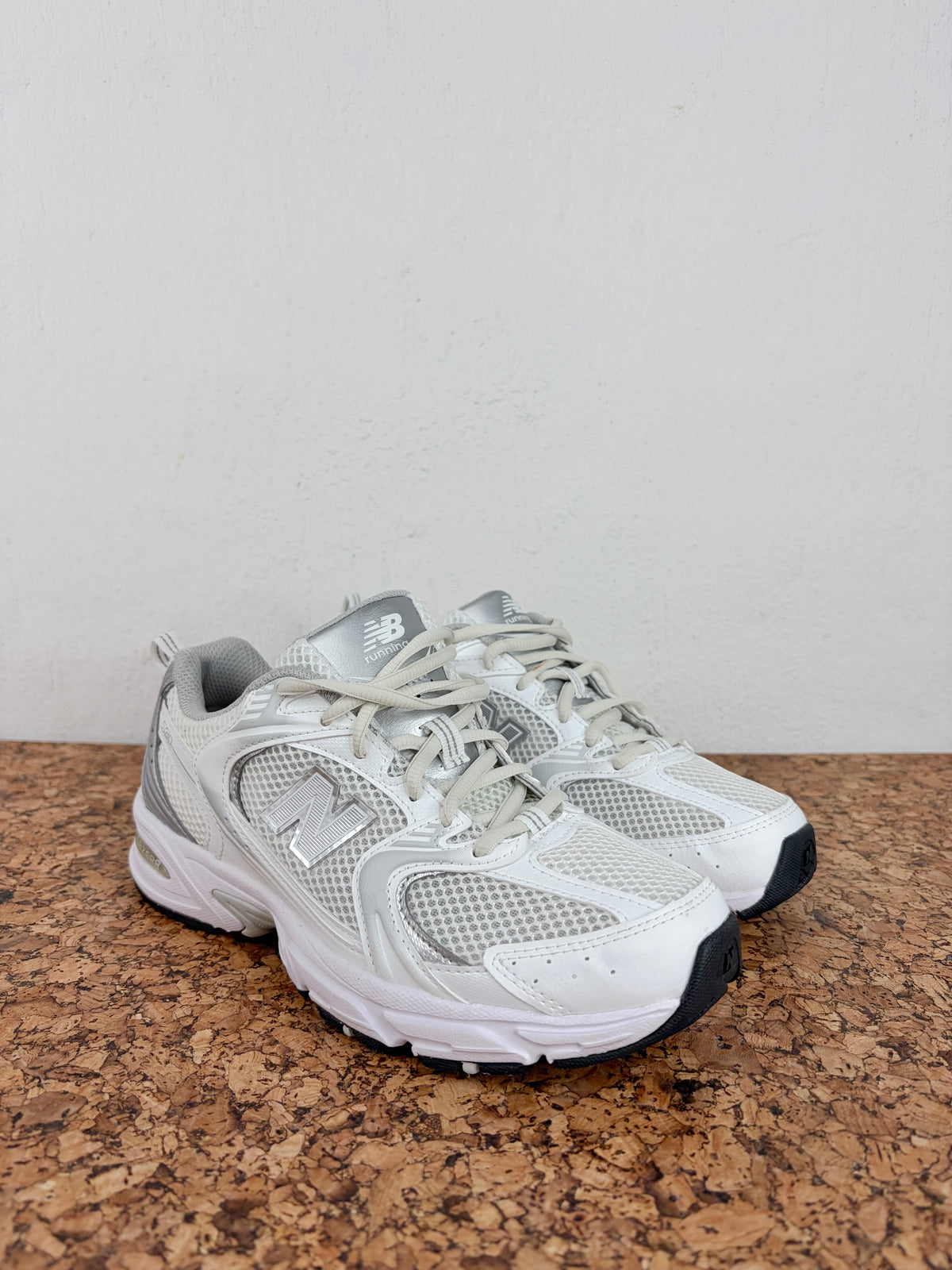 A pair of New Balance 990v4 running shoes in white and gray mesh, resting on a brown cork surface against a plain off-white wall.