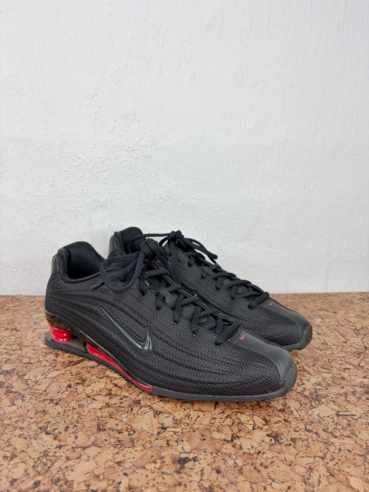 A pair of black Nike Shox sneakers with a textured knit upper, black laces, and a red Shox column visible on the side, resting on a cork surface against a white wall.