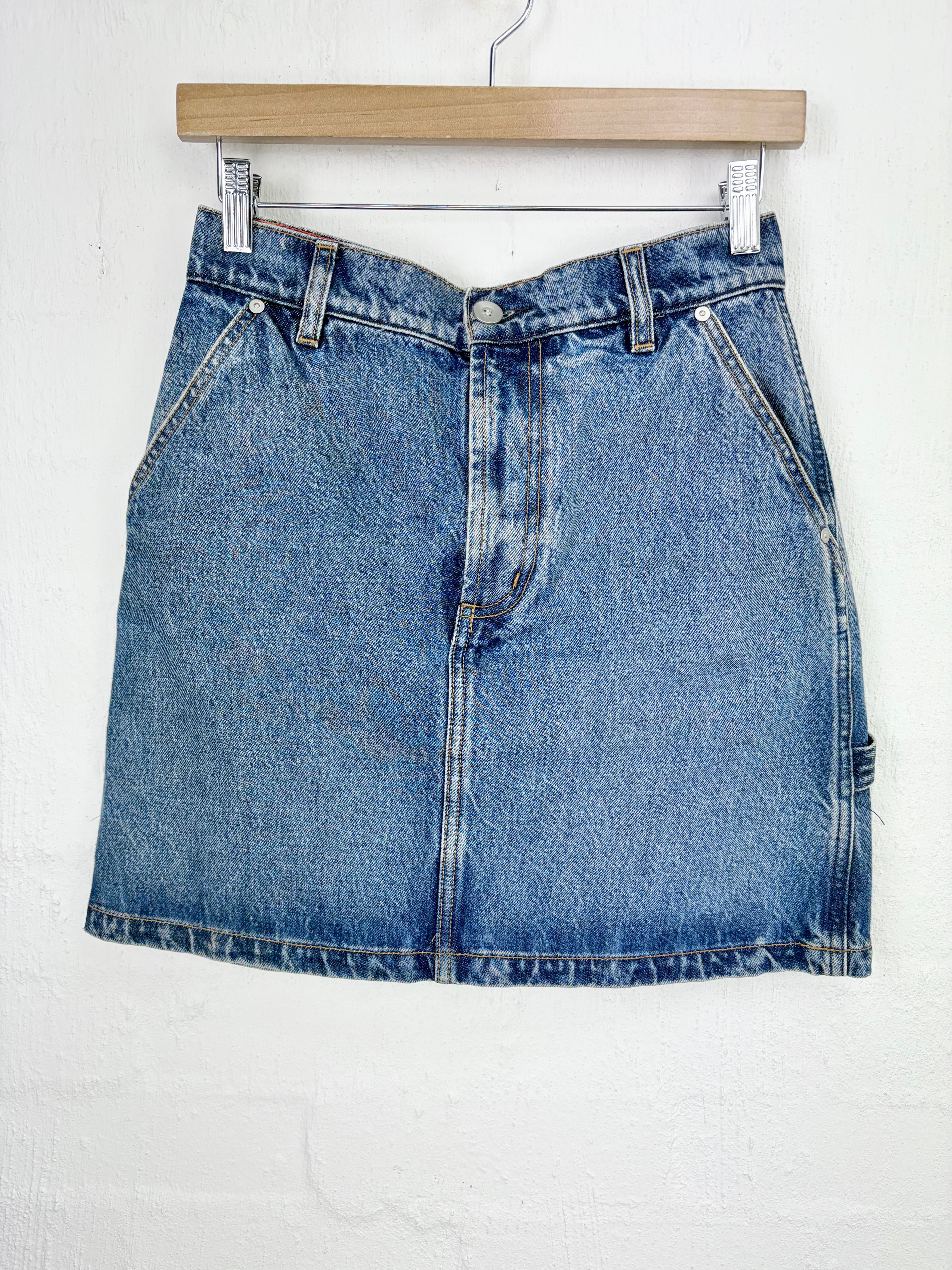 A blue denim mini skirt with a faded wash, hanging from a wooden hanger against a white textured wall.