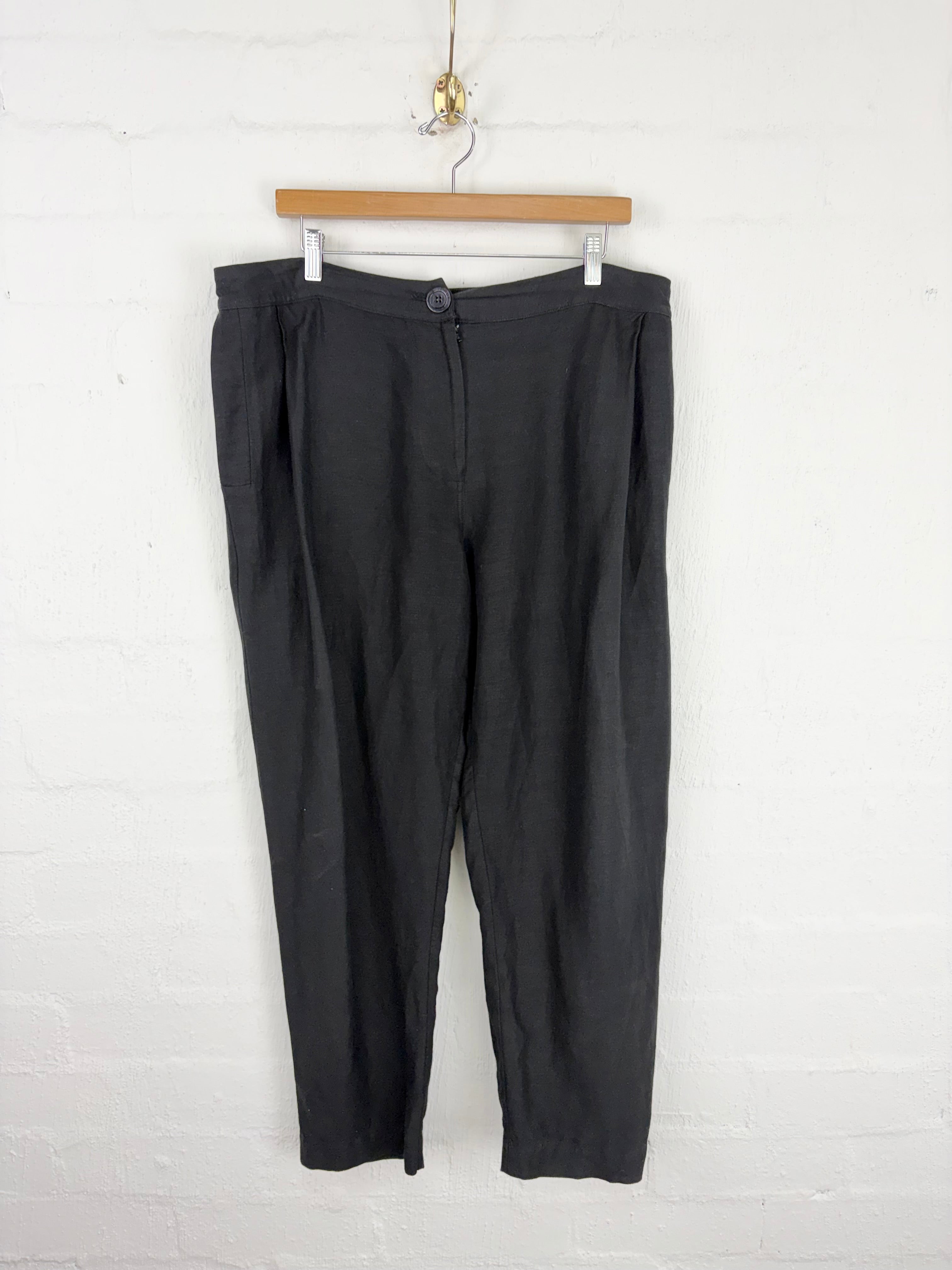 Black pleated trousers hanging from a wooden hanger against a white painted brick wall.