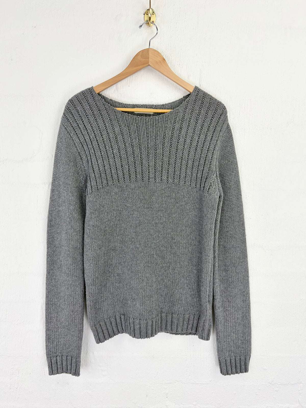 A gray knit sweater with a ribbed yoke and long sleeves, hanging on a wooden hanger against a white textured wall.