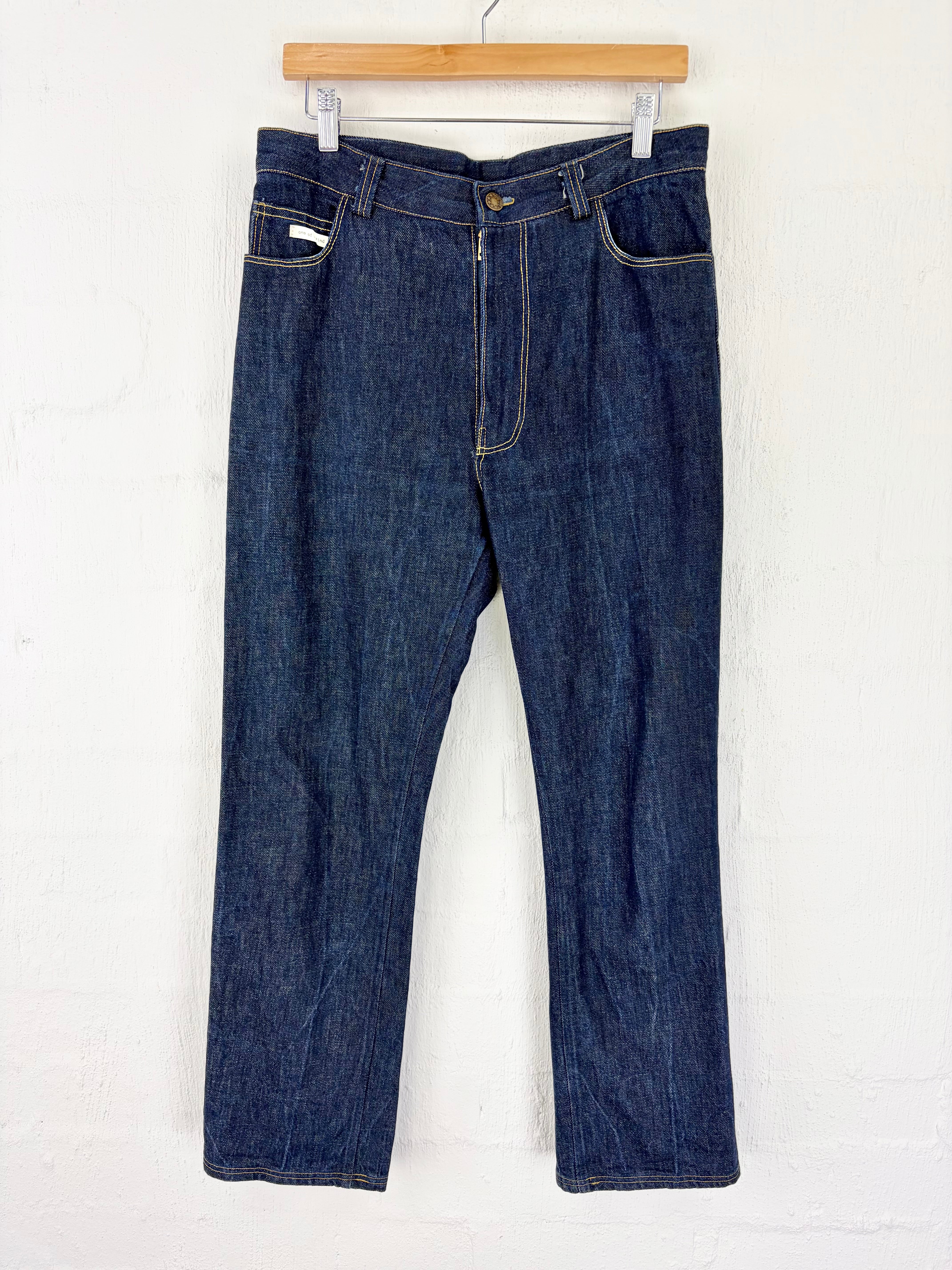 A pair of dark blue indigo denim jeans hanging from a wooden hanger against a white painted brick wall.