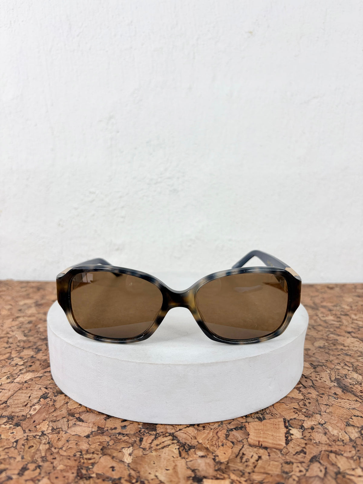 Oversized square sunglasses with a dark tortoiseshell pattern and brown lenses, displayed on a round white pedestal over a cork surface against a white wall.
