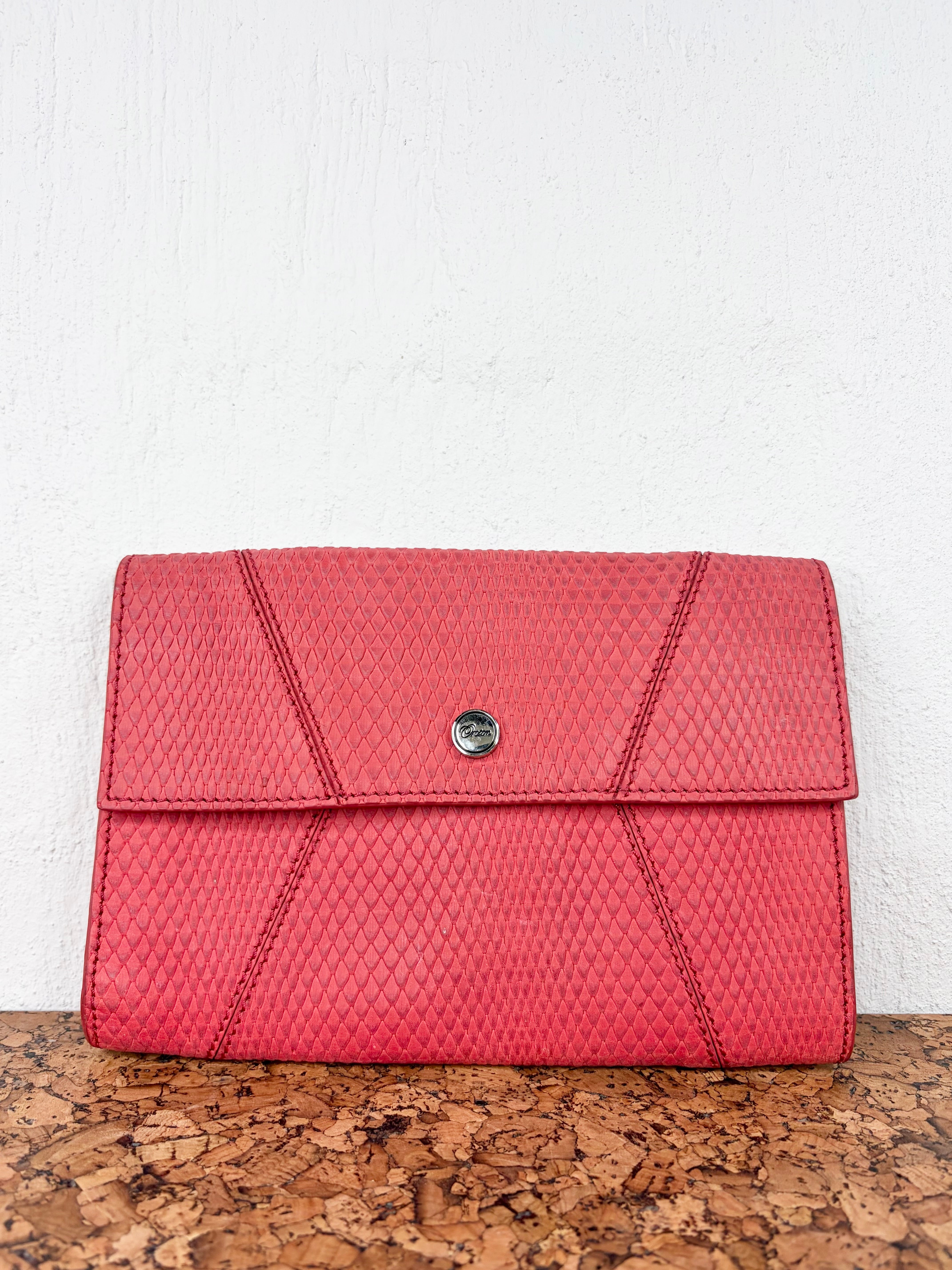 A textured red clutch bag with a small silver button closure, resting on a cork surface against a white wall.