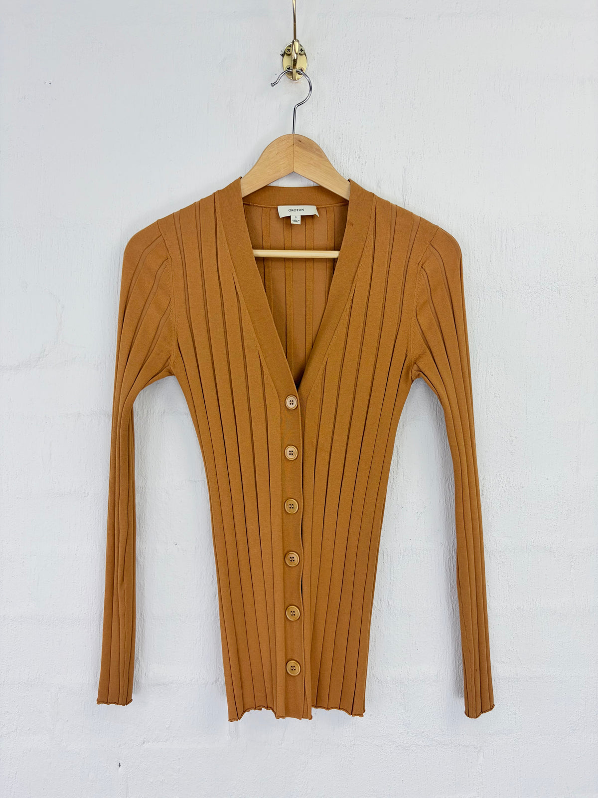 A ribbed knit cardigan sweater in a warm camel brown color, hanging on a wooden hanger against a white painted brick wall.