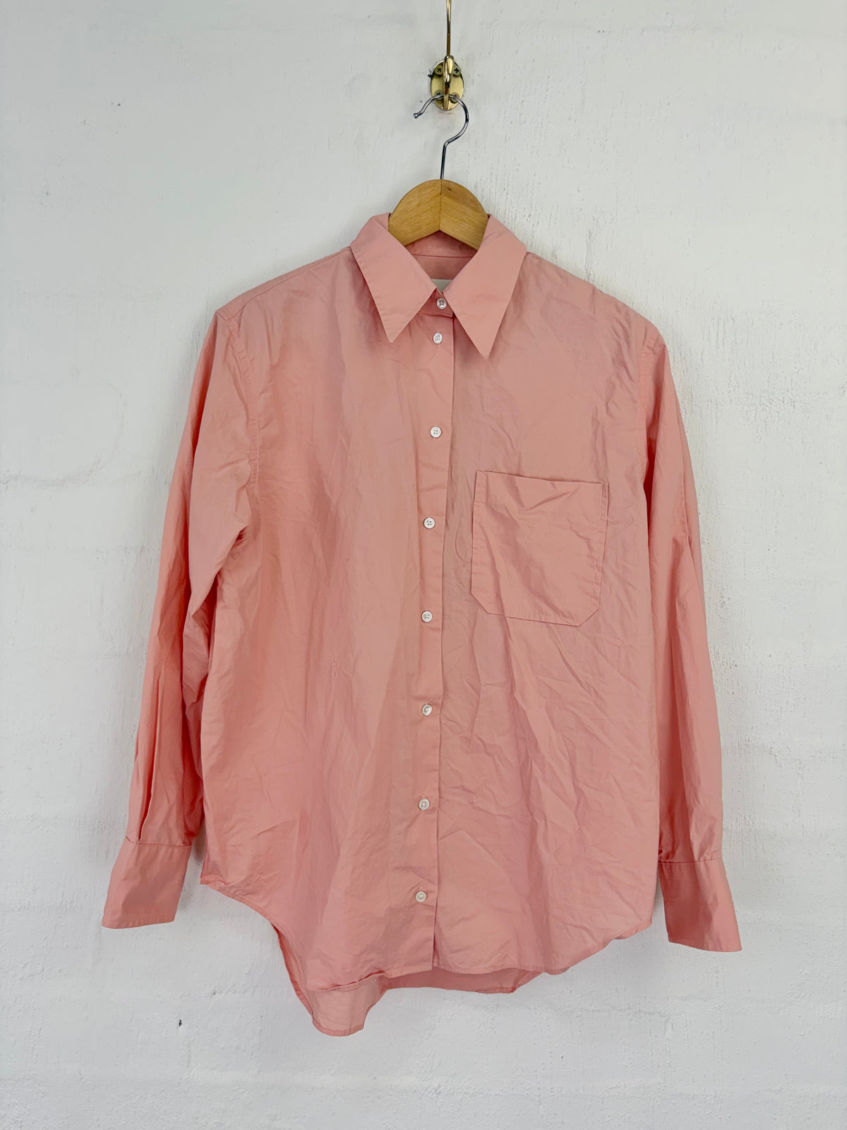 A dusty pink long-sleeve button-up shirt with a single chest pocket, hanging on a wooden hanger against a white textured wall.