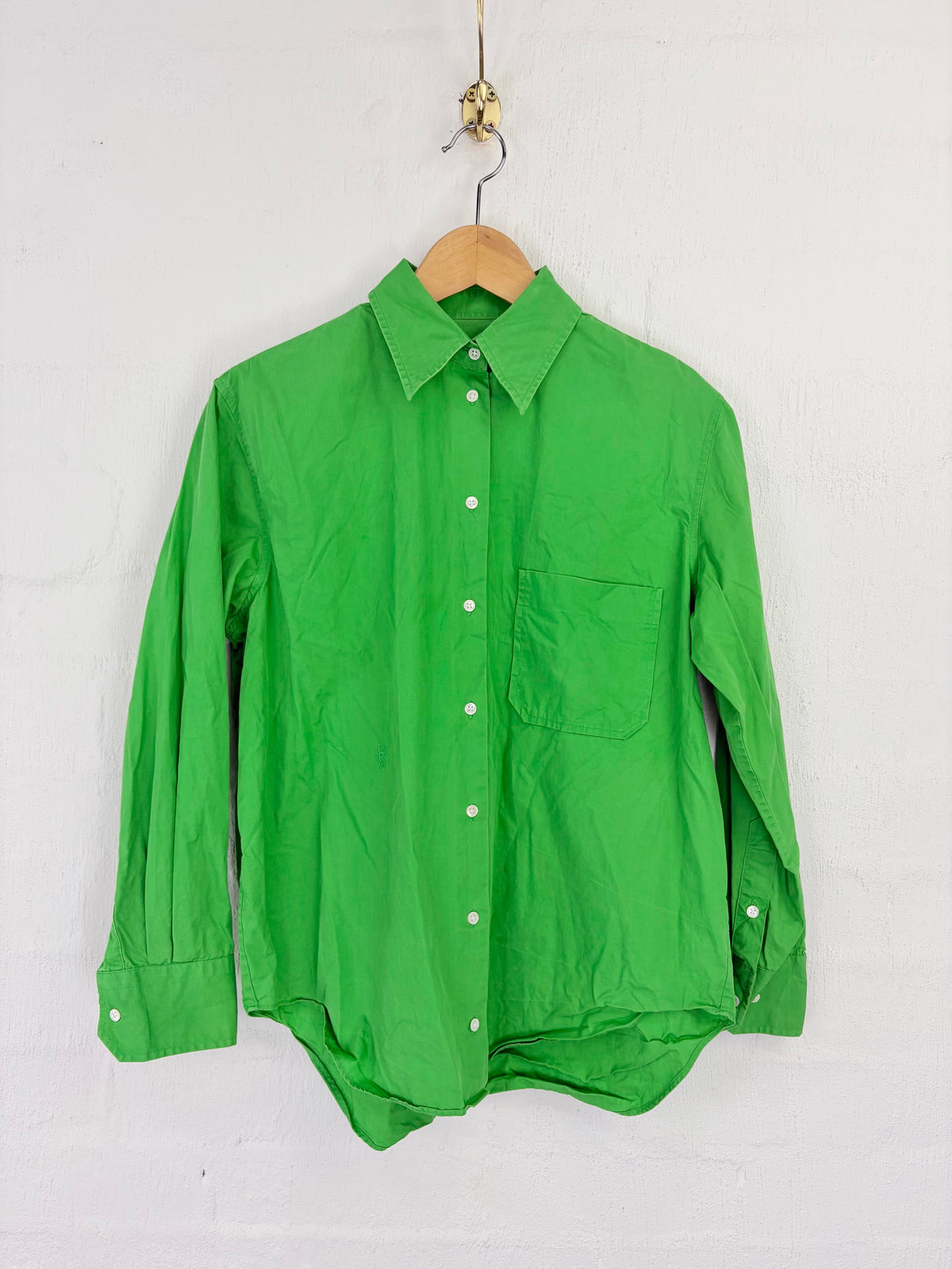 A bright green long-sleeve button-up shirt hanging on a wooden hanger against a white painted brick wall.