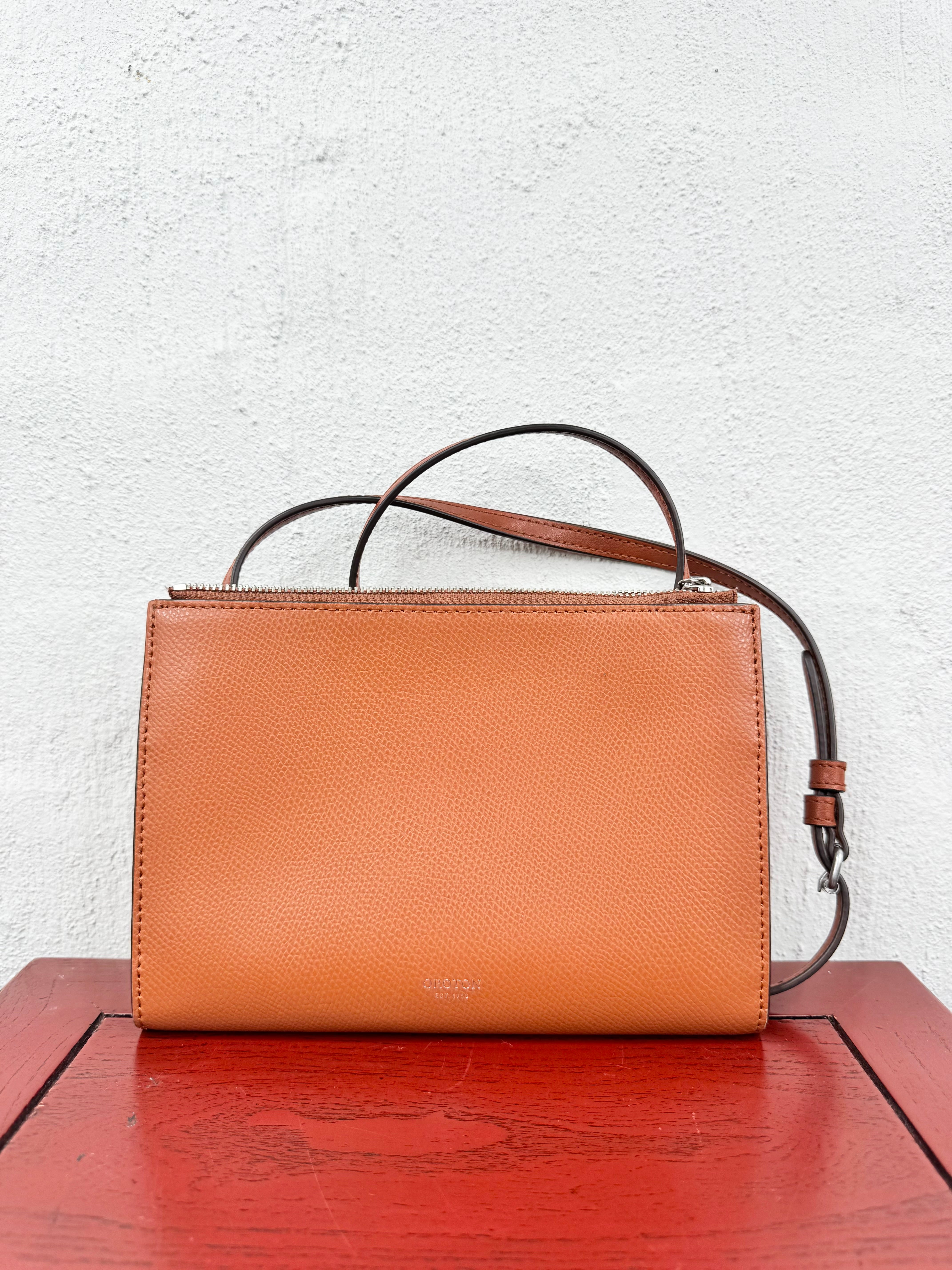 A tan pebbled leather handbag with a top zipper, double top handle, and detachable shoulder strap, displayed on a red wooden table against a white textured wall.