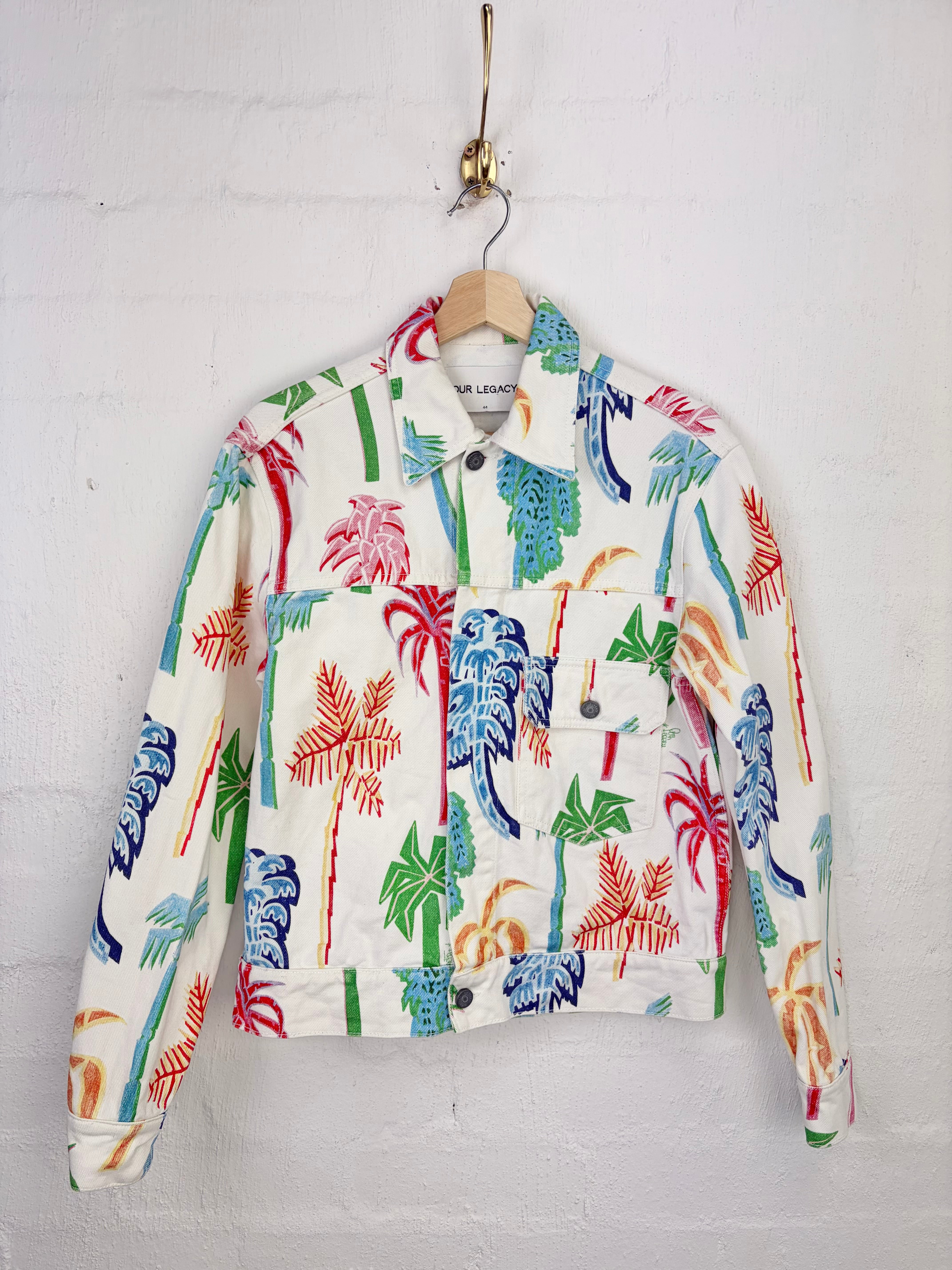 A white denim jacket with a colorful palm tree print, hanging on a wooden hanger against a white brick wall.