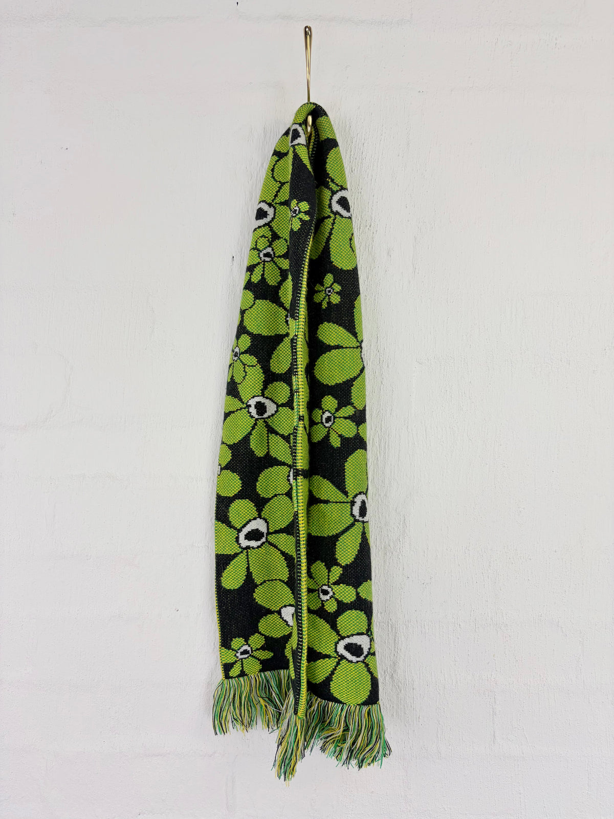 A black knit scarf with a vibrant green floral pattern and multicolor fringe, hanging from a metal hook against a white painted brick wall.