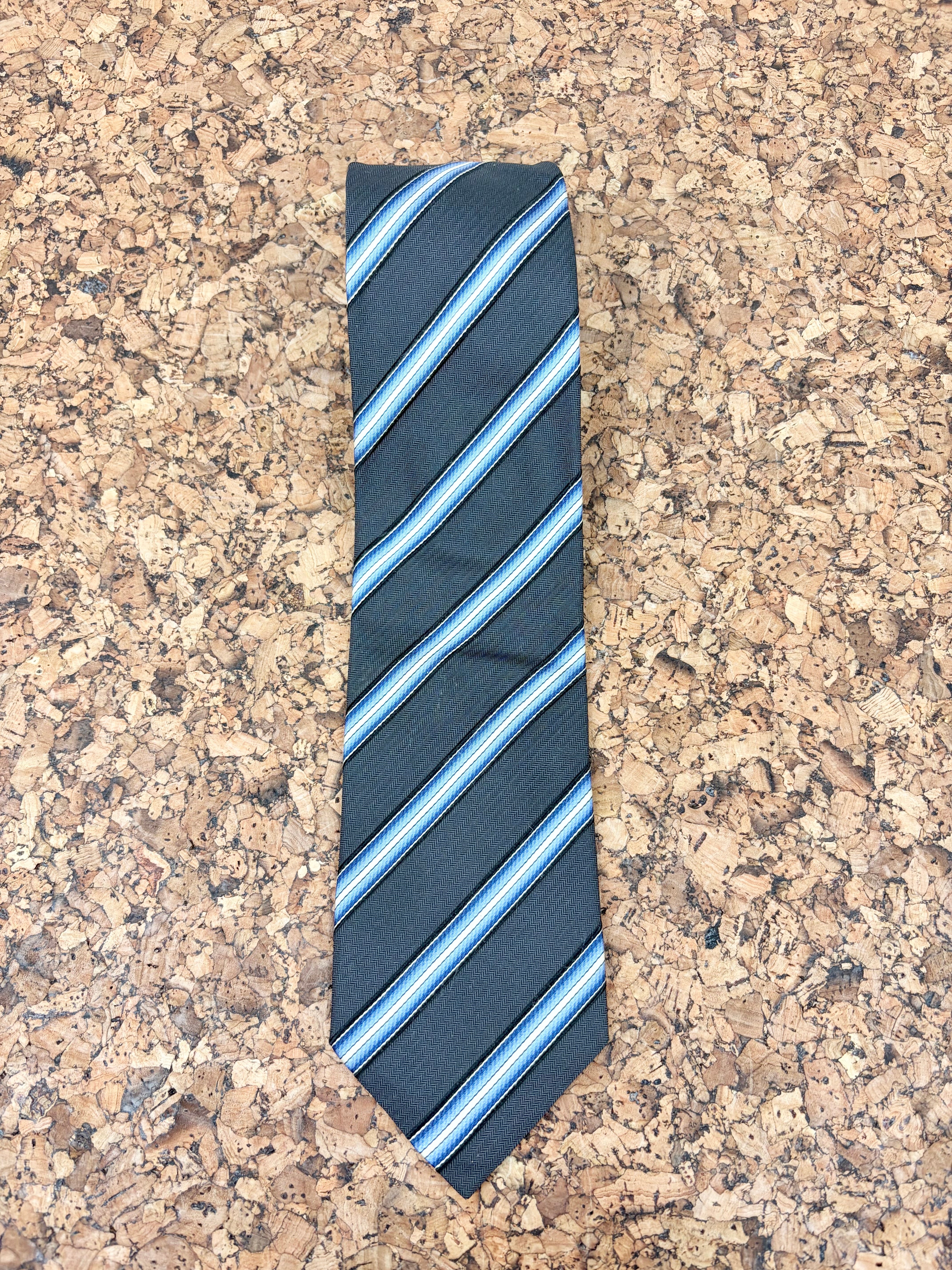 A dark navy blue necktie with diagonal stripes in light blue, white, and thin red lines, laid flat on a textured cork surface.