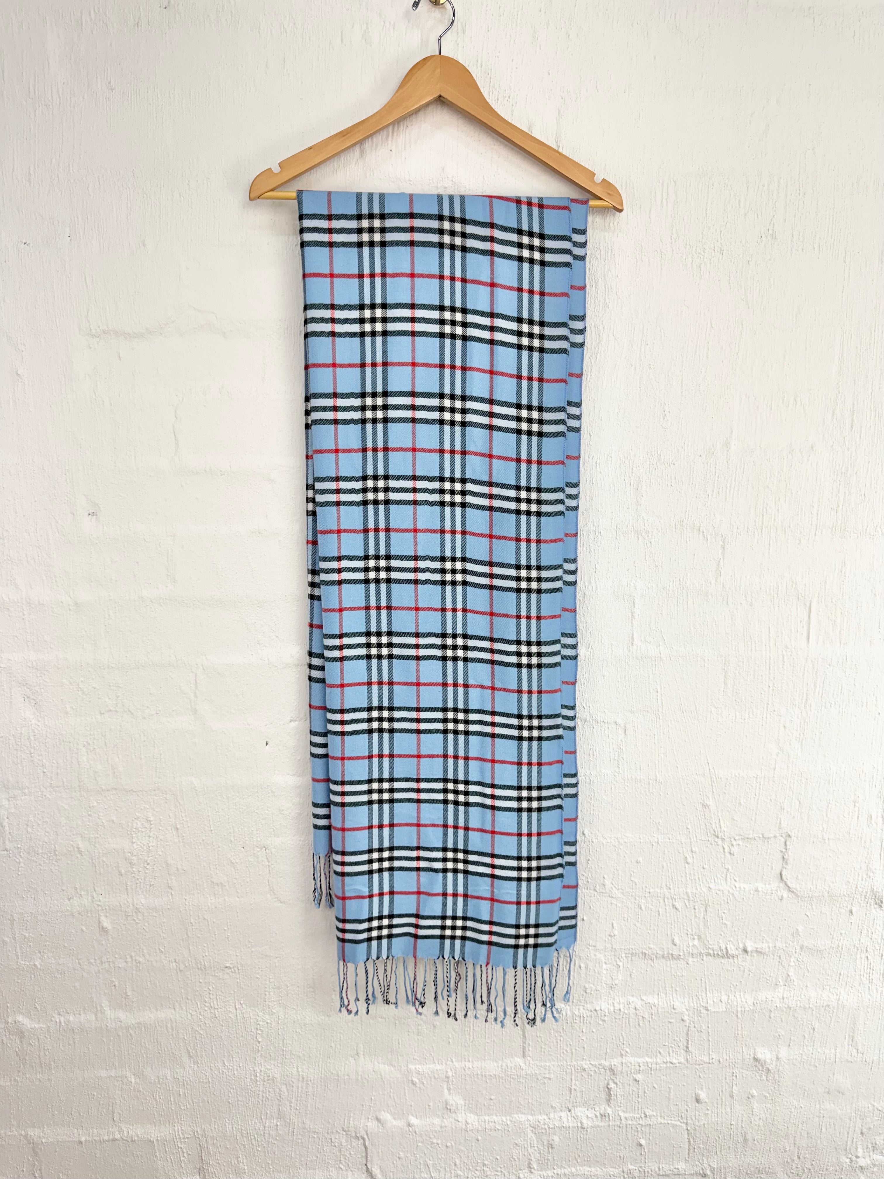 A light blue plaid scarf with black, white, and red stripes, hanging from a wooden hanger against a white painted brick wall.