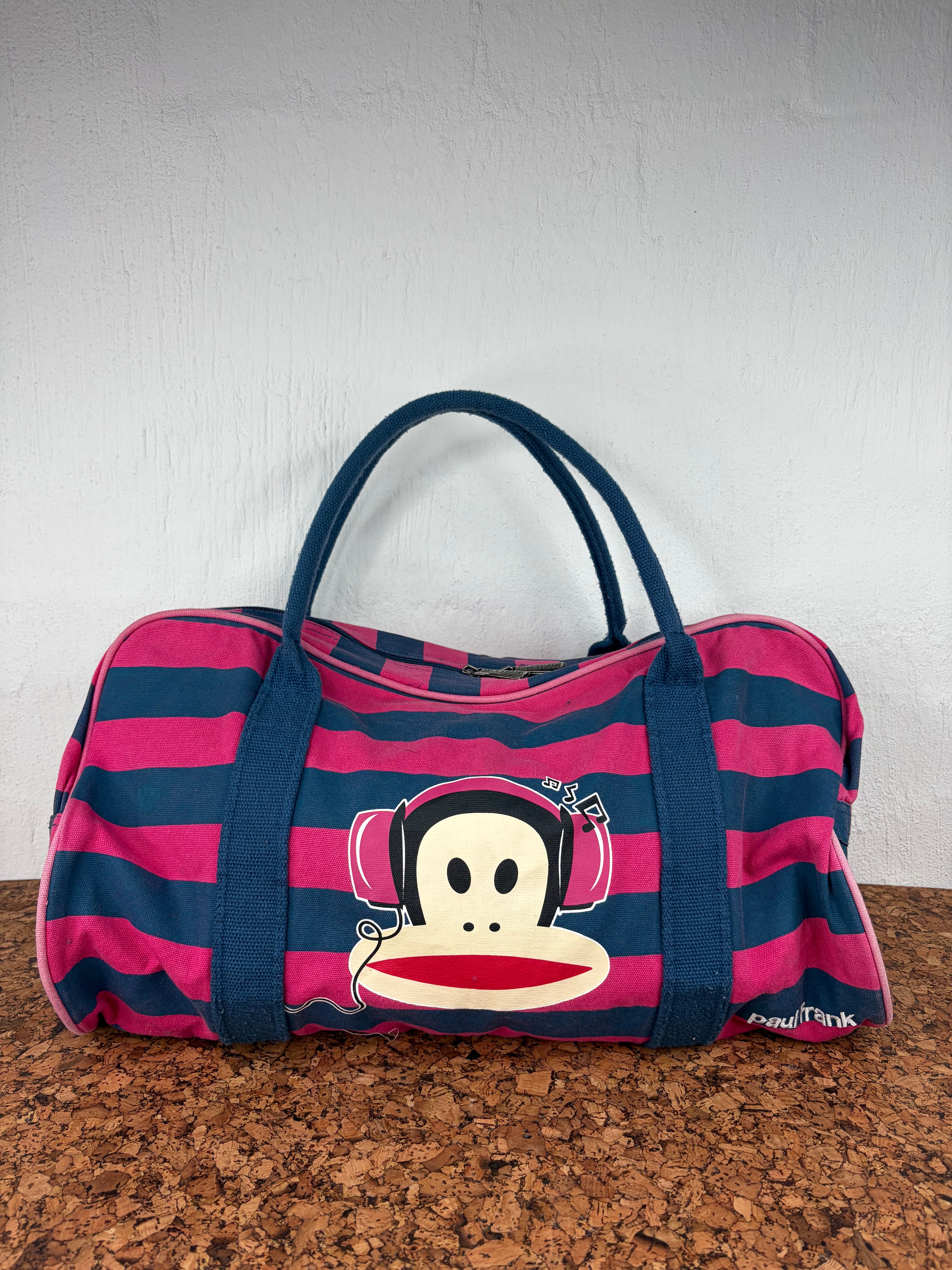 A pink and navy blue striped duffle bag with a large graphic of the San Paulo sock monkey wearing headphones on the front, set on a brown cork surface against a white wall.