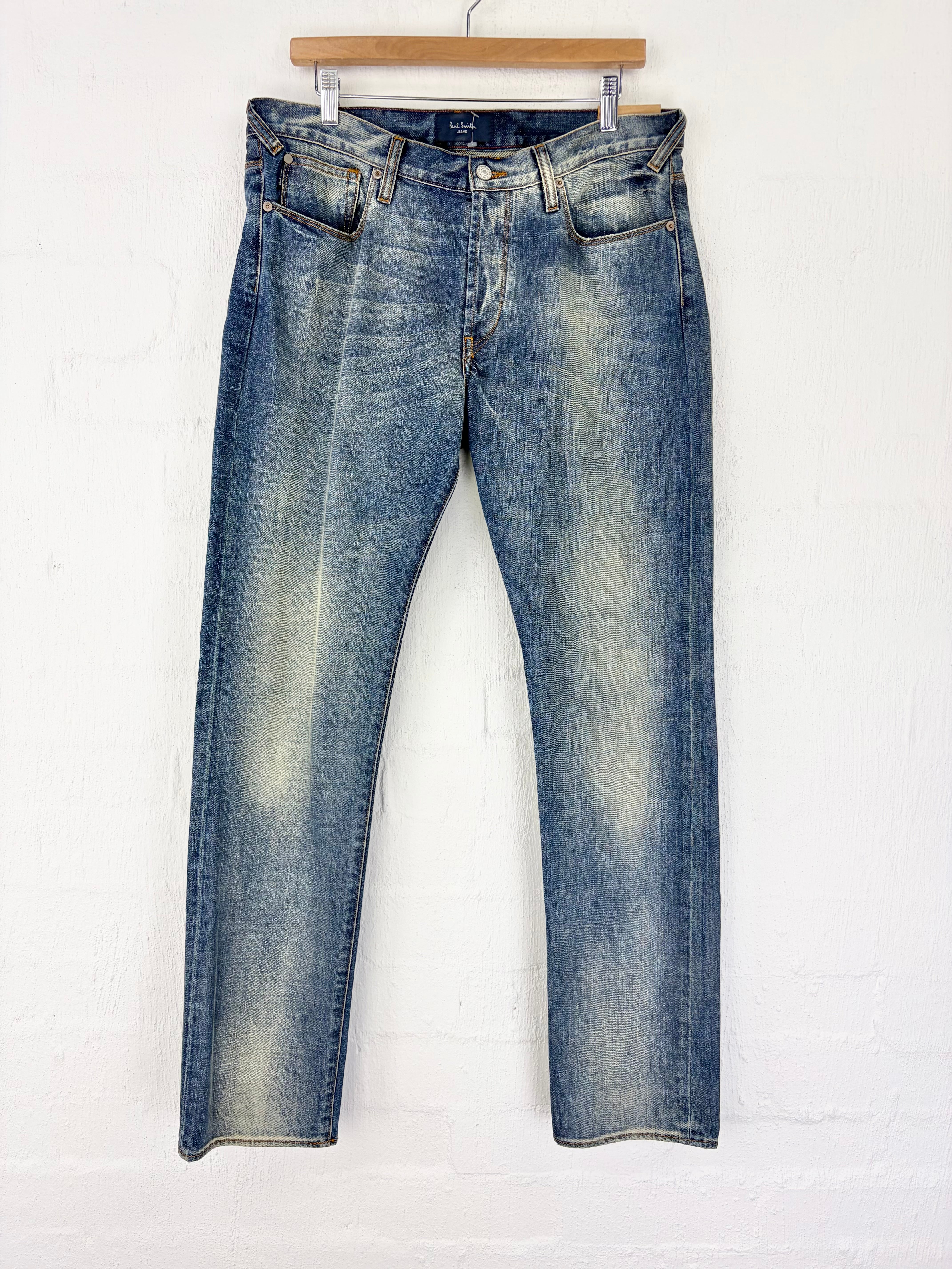 A pair of medium-wash blue denim jeans with a faded, distressed finish, hanging from a wooden hanger against a white painted brick wall.
