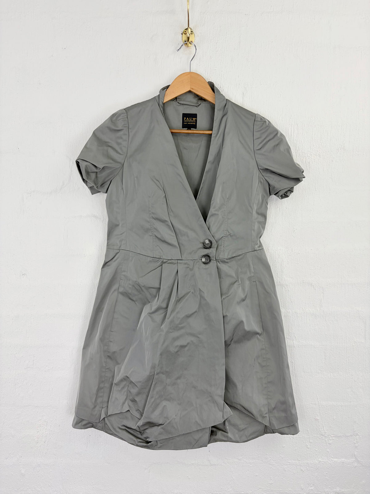 A gray short-sleeve wrap dress with a shawl collar and button front, hanging on a wooden hanger against a white brick wall.
